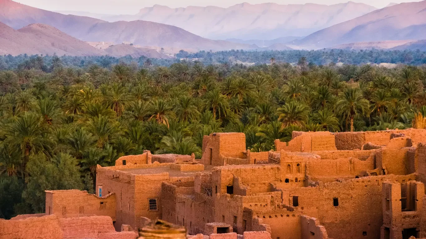 Ancient mud-brick village with multiple structures, set against a lush green date palm oasis and mountain range in the background.