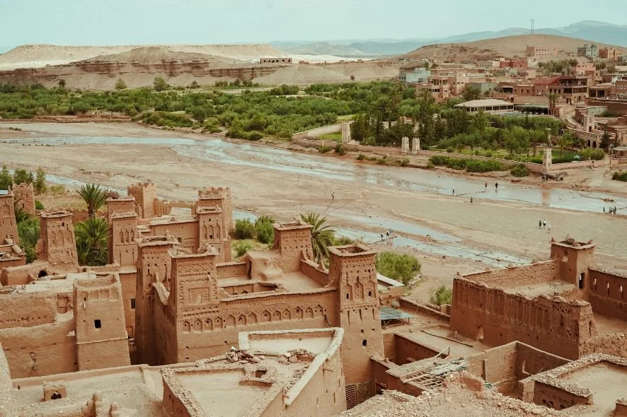 Historic mud-brick buildings near a river with green vegetation, set against a hilly landscape in a desert region.