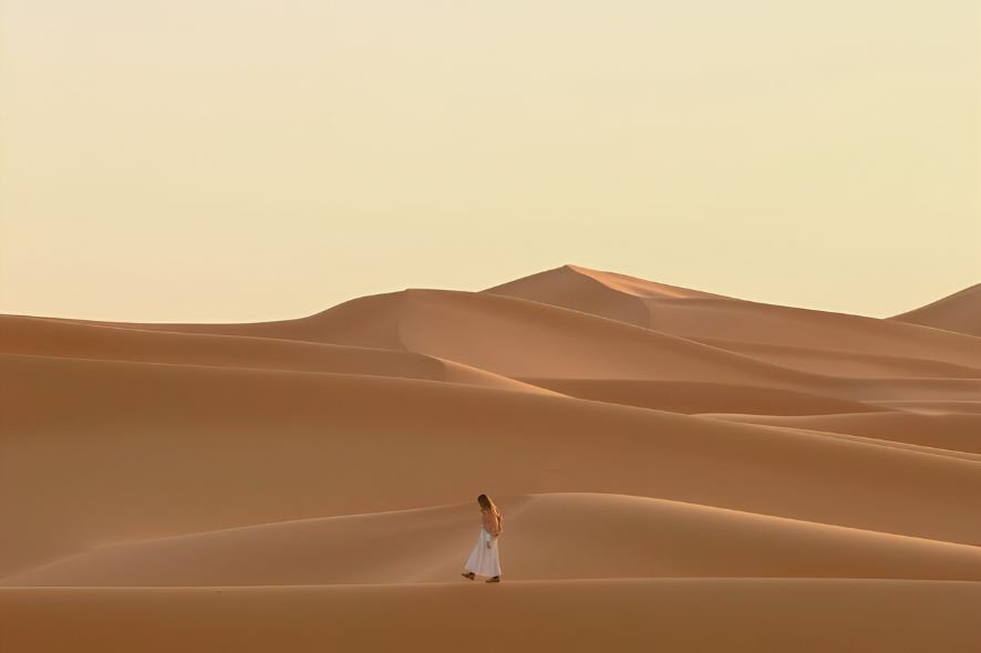 A woman in a white dress walking on sand dunes in a desert during sunset.