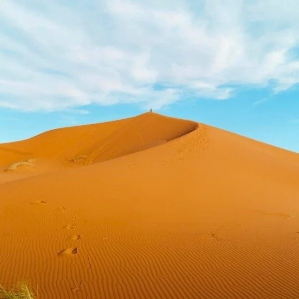 Footsteps leading up a sandy desert dune with a person at the top against a blue sky with clouds.