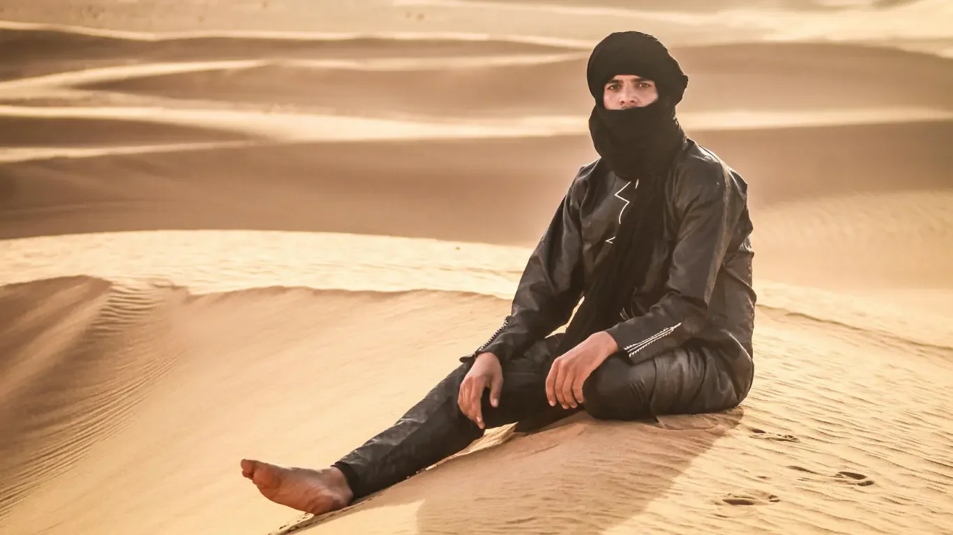 A person sitting on sand dunes in a desert, wearing a black turban and face covering, dressed in black leather clothing.
