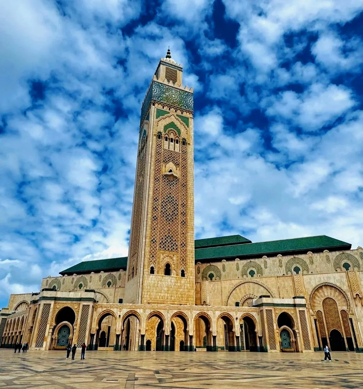 The Hassan II Mosque in Casablanca, Morocco, featuring a tall minaret with intricate designs and green roofs, set against a partly cloudy sky.