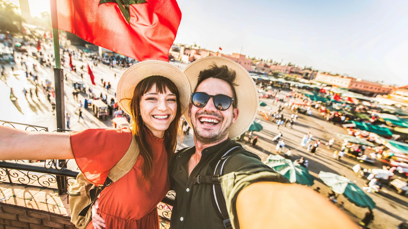 A smiling man and woman taking a selfie together outdoors in a busy marketplace, both wearing wide-brimmed hats and casual summer clothing, with the woman dressed in a red top and the man in black sunglasses and a black shirt.