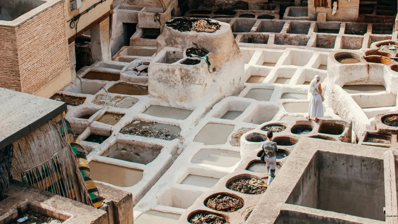 Two women walking through a traditional Moroccan tannery with numerous stone dyeing pits filled with colored liquids.