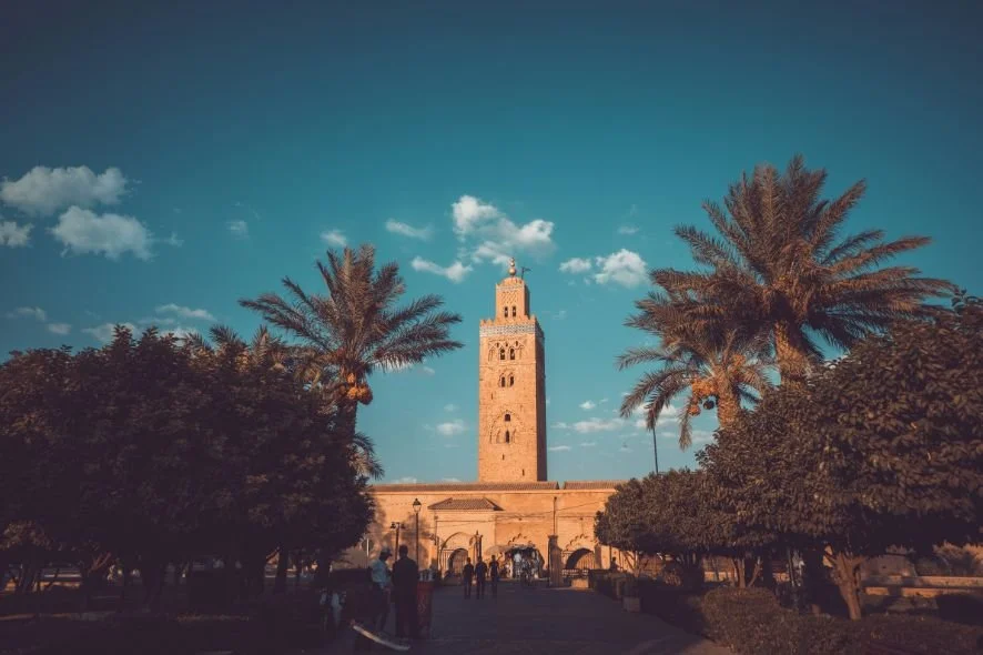 Historic Moroccan city square with tall minaret cathedral, palm trees, and people walking in the foreground under a blue sky with clouds.