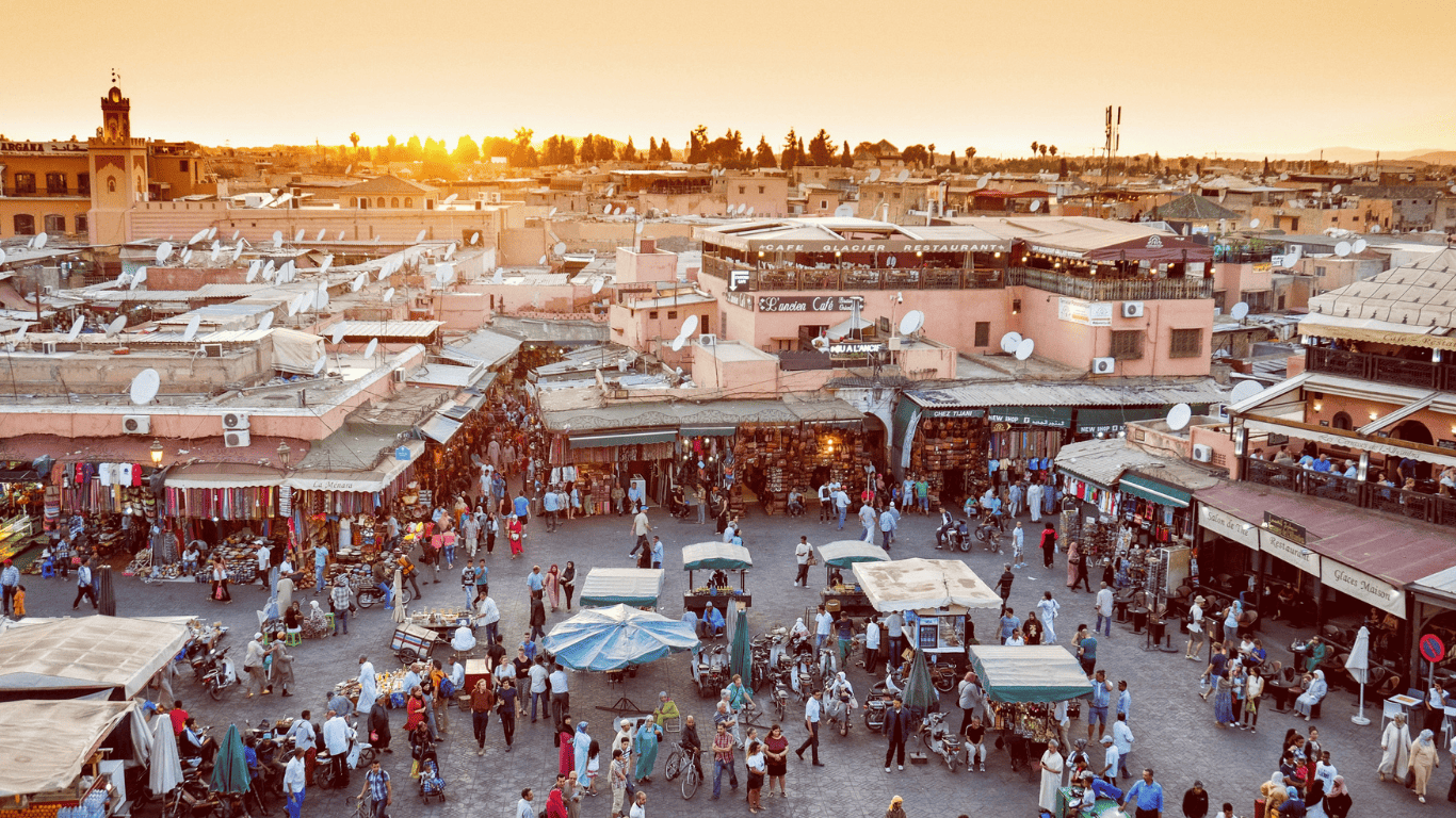 A busy marketplace in a city during sunset, with many people shopping and walking among vendor stalls and street vendors.