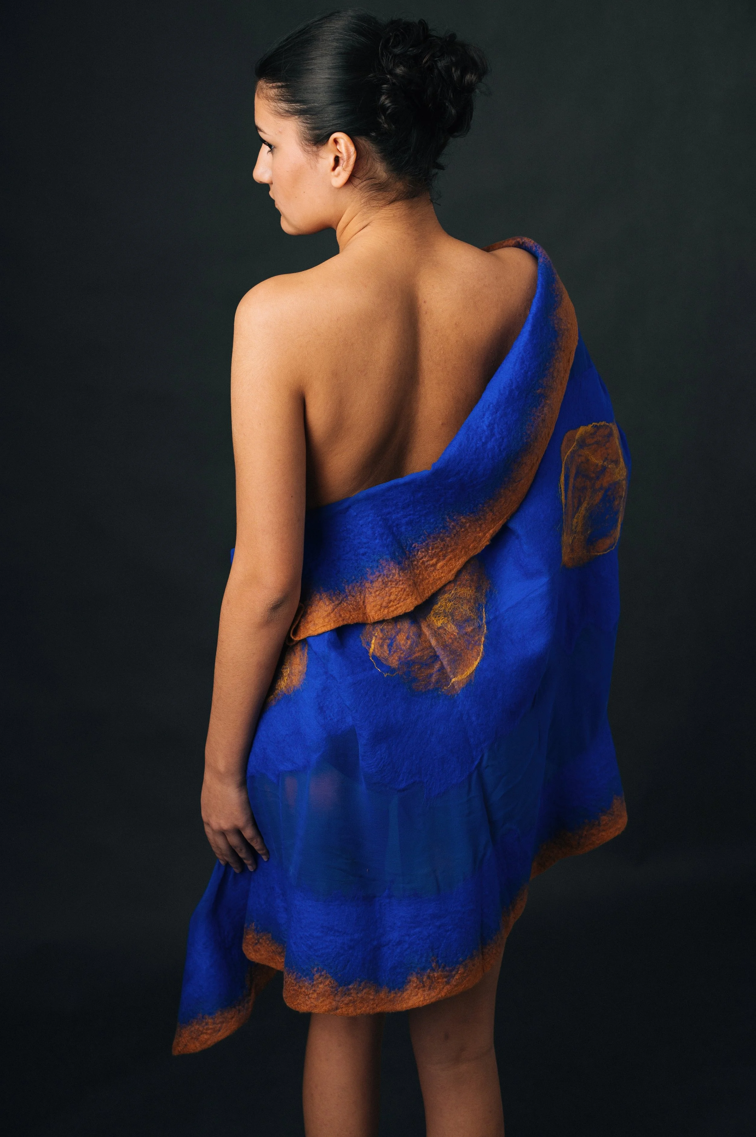 A woman with dark hair styled up, facing away, partially wrapped in a blue and orange shawl, standing against a dark background.