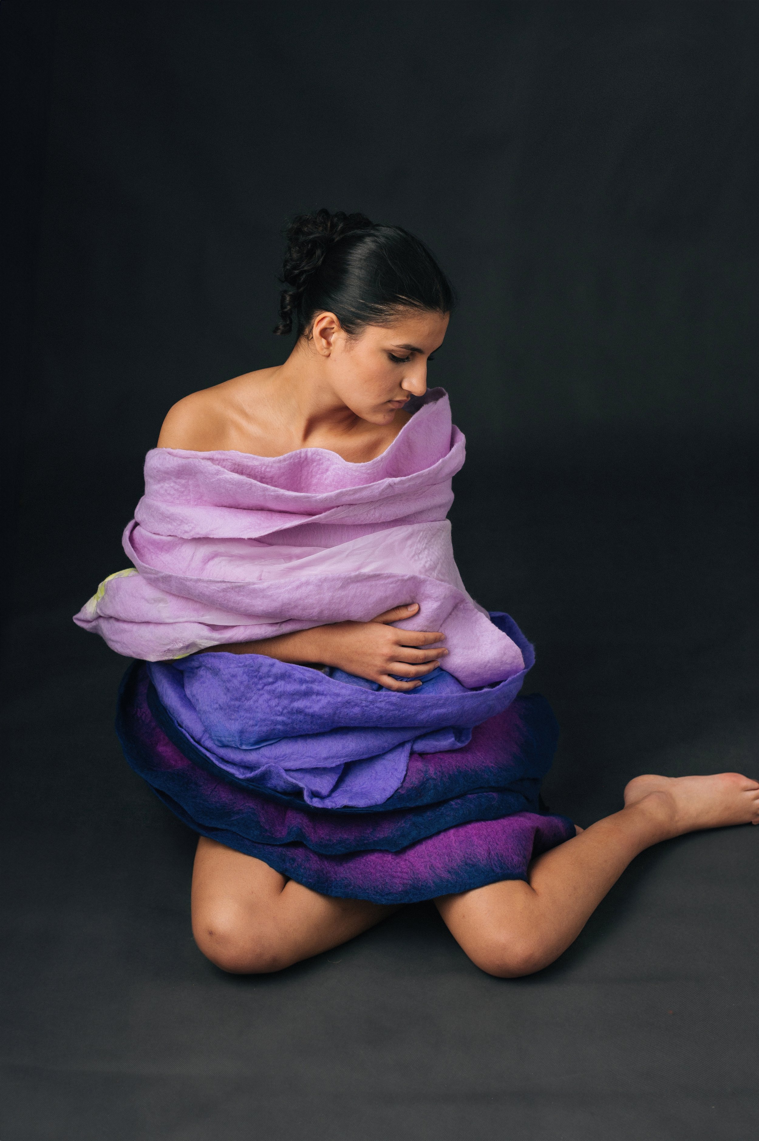 A woman sitting on the floor, wrapped in layered, colorful fabric resembling a mountain landscape, with a dark background.