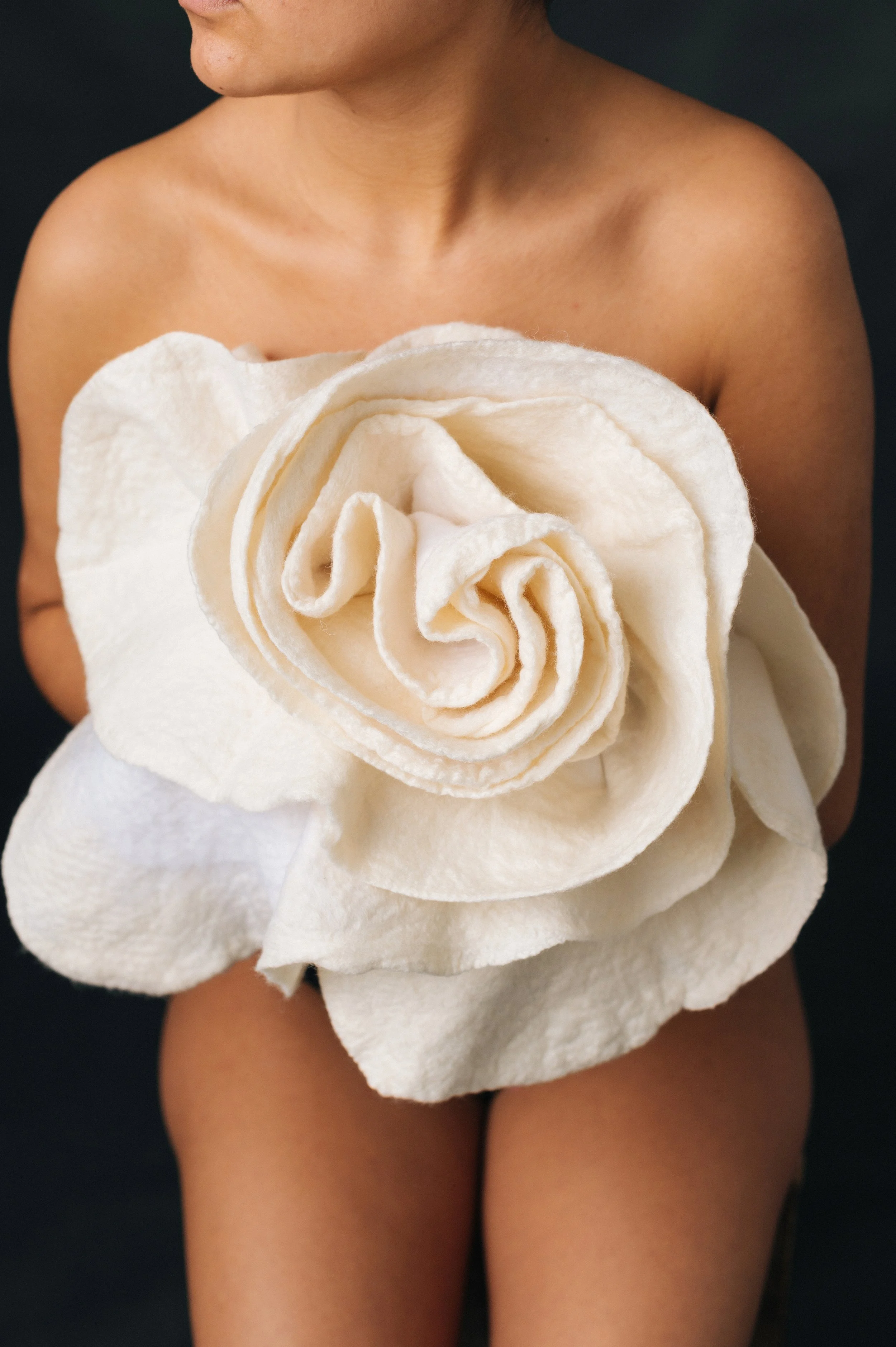 Person holding a large, white, fabric flower with multiple layers, standing nude against a black background.