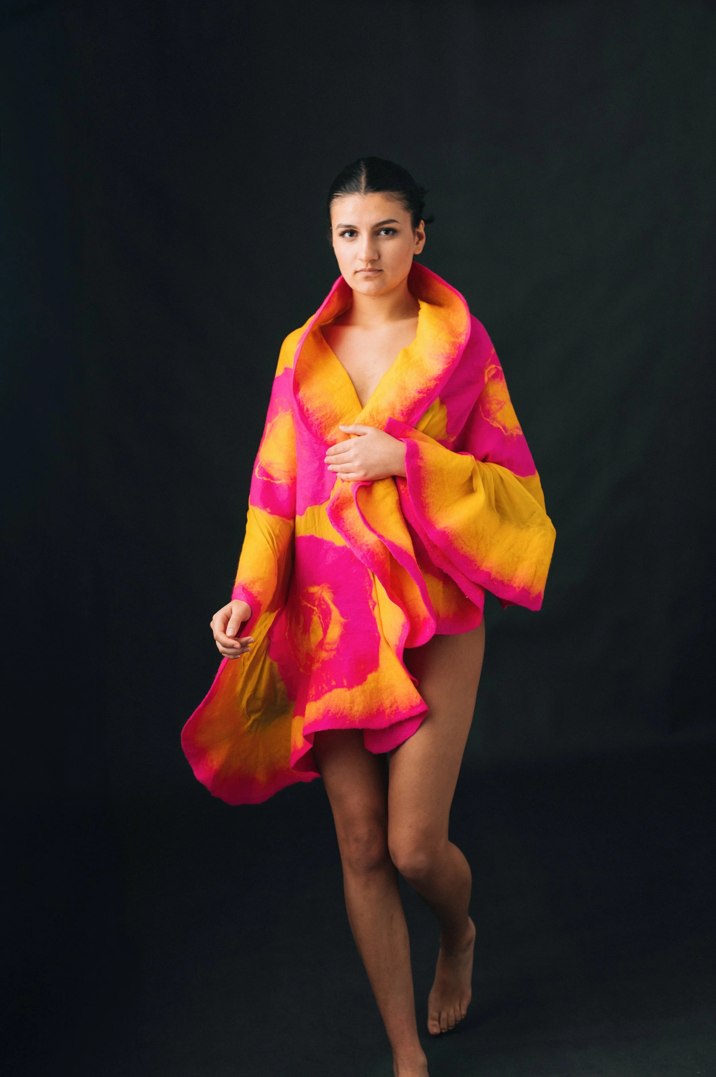 A woman with dark hair and fair skin, partially draped in a colorful yellow and pink patterned fabric, standing against a dark background.