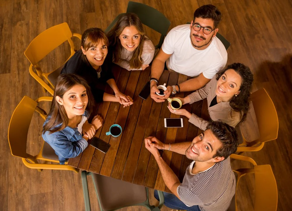Six young adults sitting around a wooden table, smiling, with cups of coffee and smartphones on the table, viewed from above.