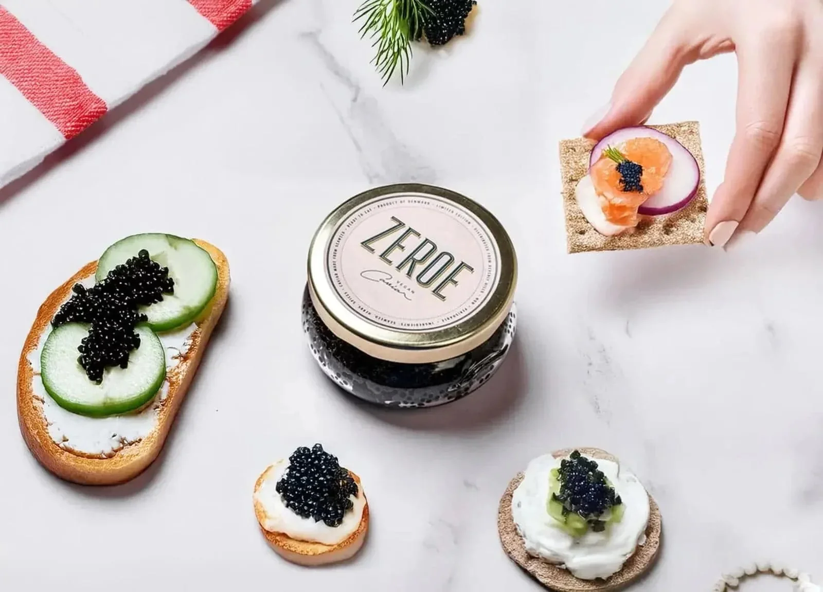 Assorted appetizers with caviar on crackers and bread slices, a jar of 'Zeroe' caviar, garnished with cucumber, radish, and dill on a marble surface.