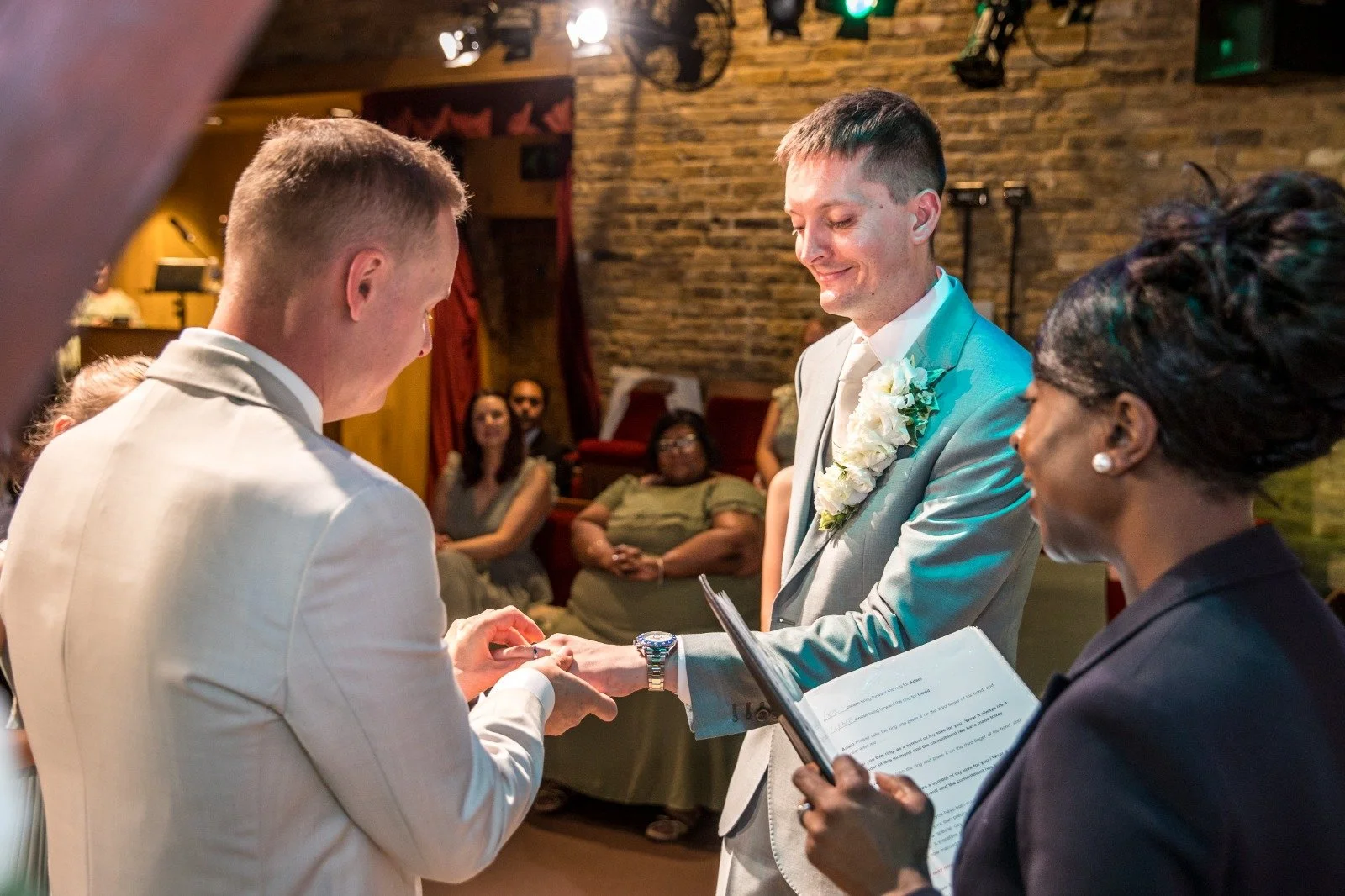 Two men, smiling at one another during their wedding ceremony. One groom is sliding the ring onto the other grooms finger at their wedding in a theatre. They are both wearing light suits.