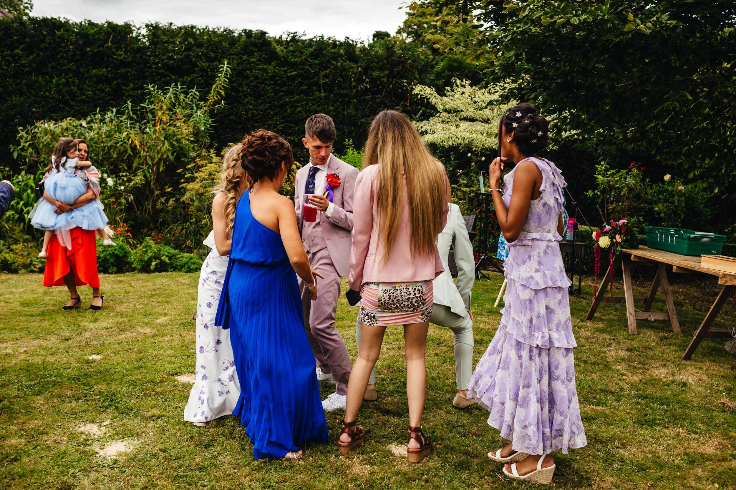 Group of people dancing outdoors at a wedding drinks reception  at a wedding in Wales, as Emma Katherine Singer sings, dressed in colourful formal attire, with a woman holding a child in the background.