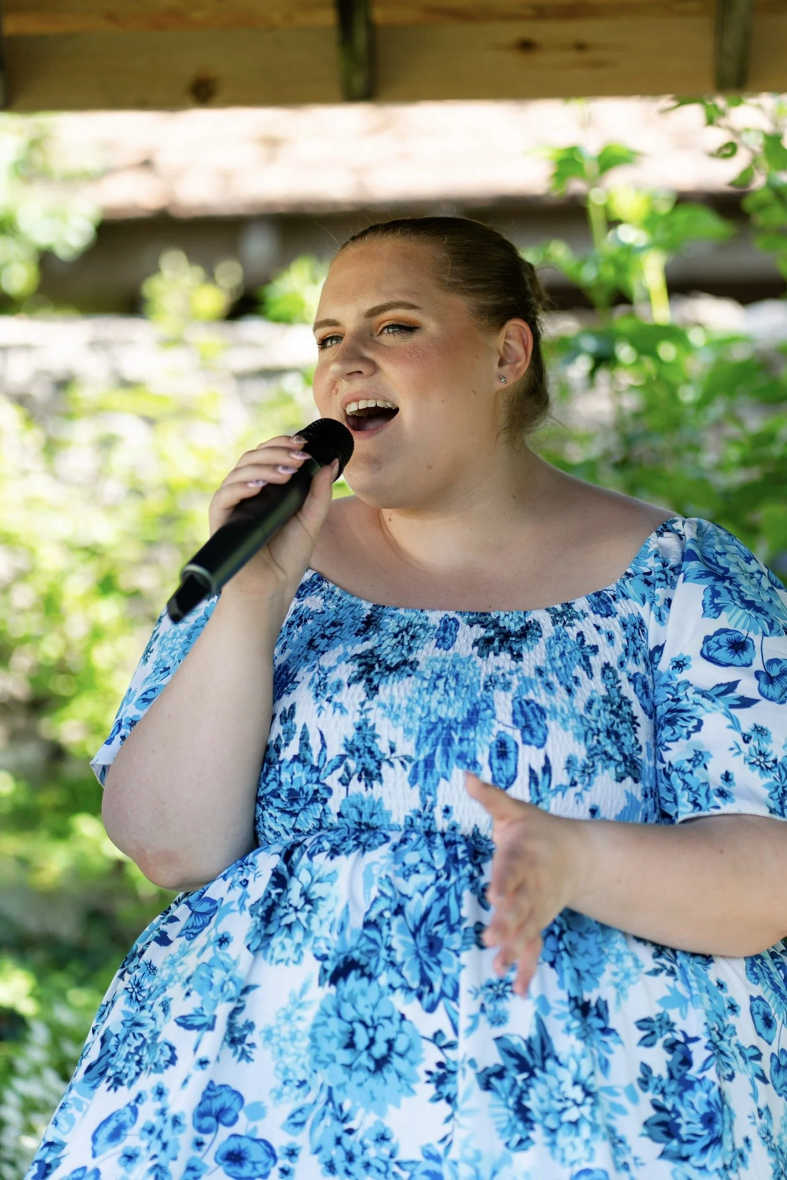 Emma Katherine Singer, Gloucestershire based wedding singer, singing into a microphone outdoors at The Great Tythe Barn, Tetbury, wearing a blue and white floral dress, with green foliage in the background.