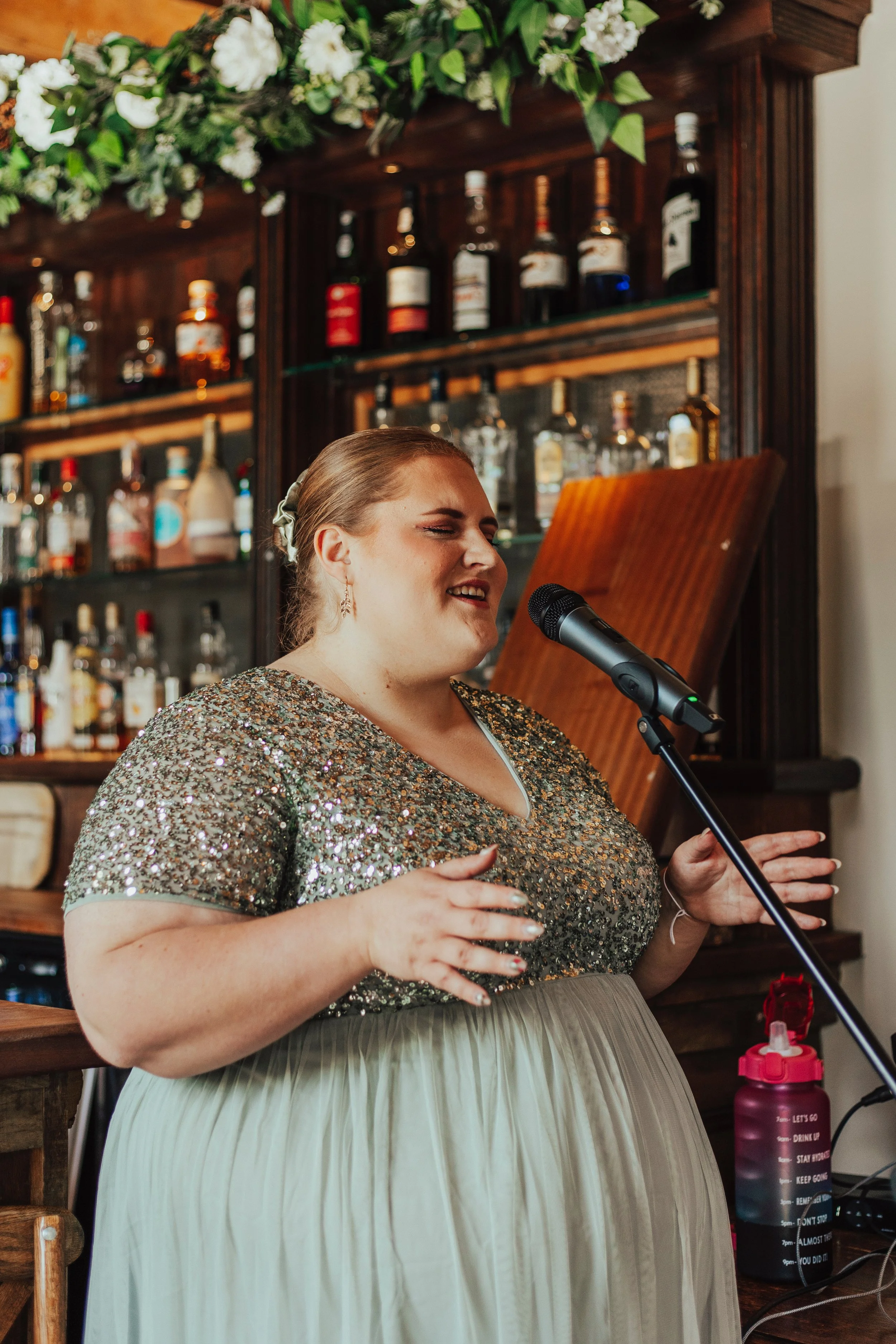 Emma Katherine Singer, a Gloucestershire based wedding singer, singing at a microphone for a wedding ceremony in Bristol, wearing a green dress with a glittery top at the back of a wedding ceremony.