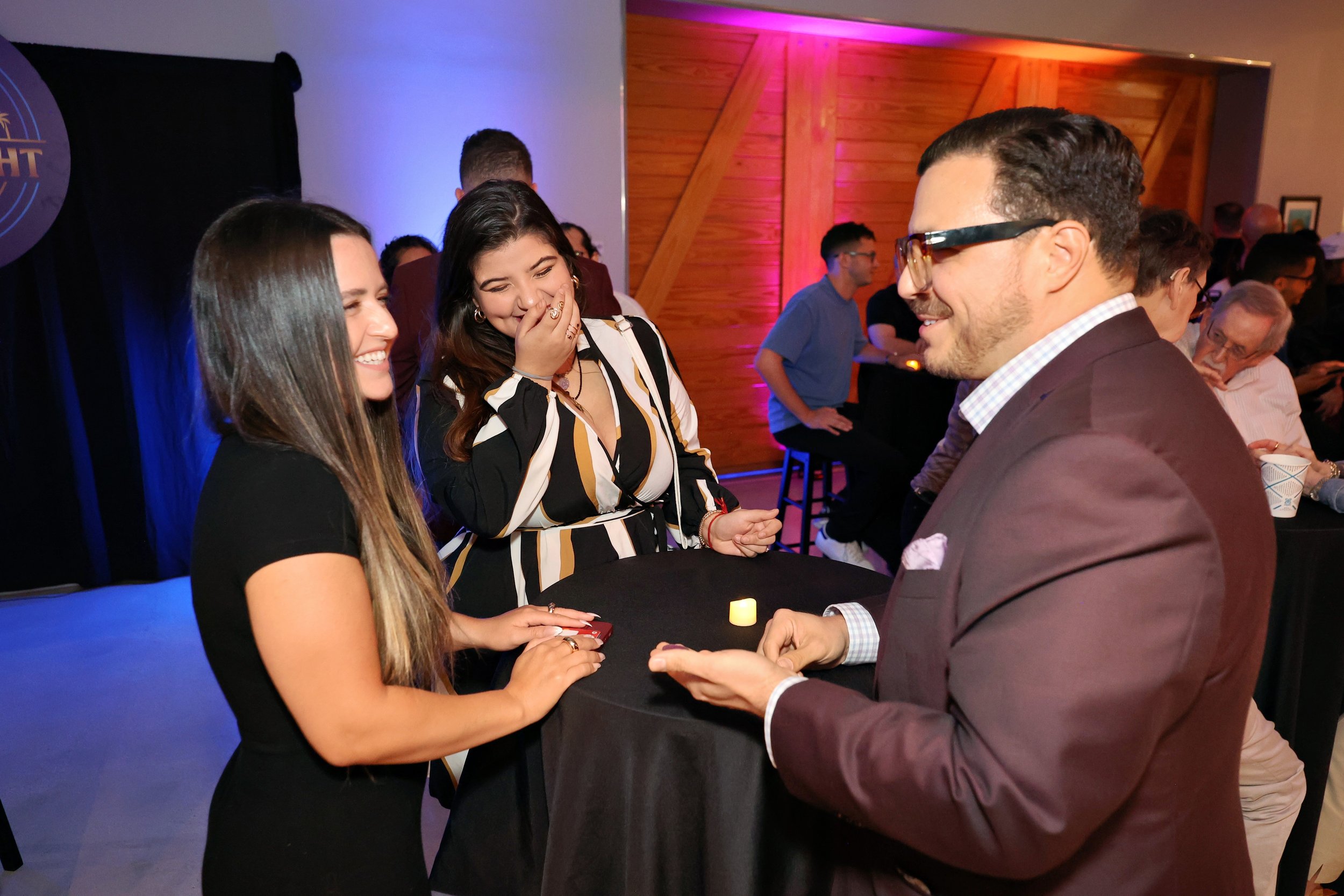 Michael Andrew performing close-up magic for two guests at a Miami event as they laugh and react.
