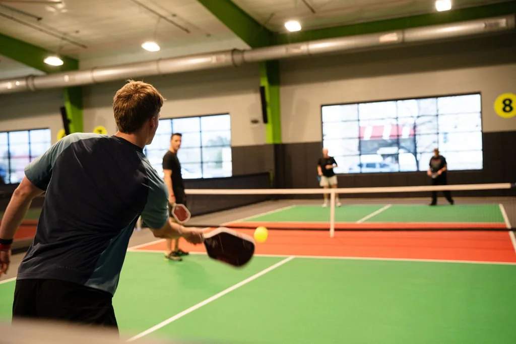 Four people play pickleball on a green court in an indoor pickleball facility. One player is seen from behind, while the others are in the background facing the camera.