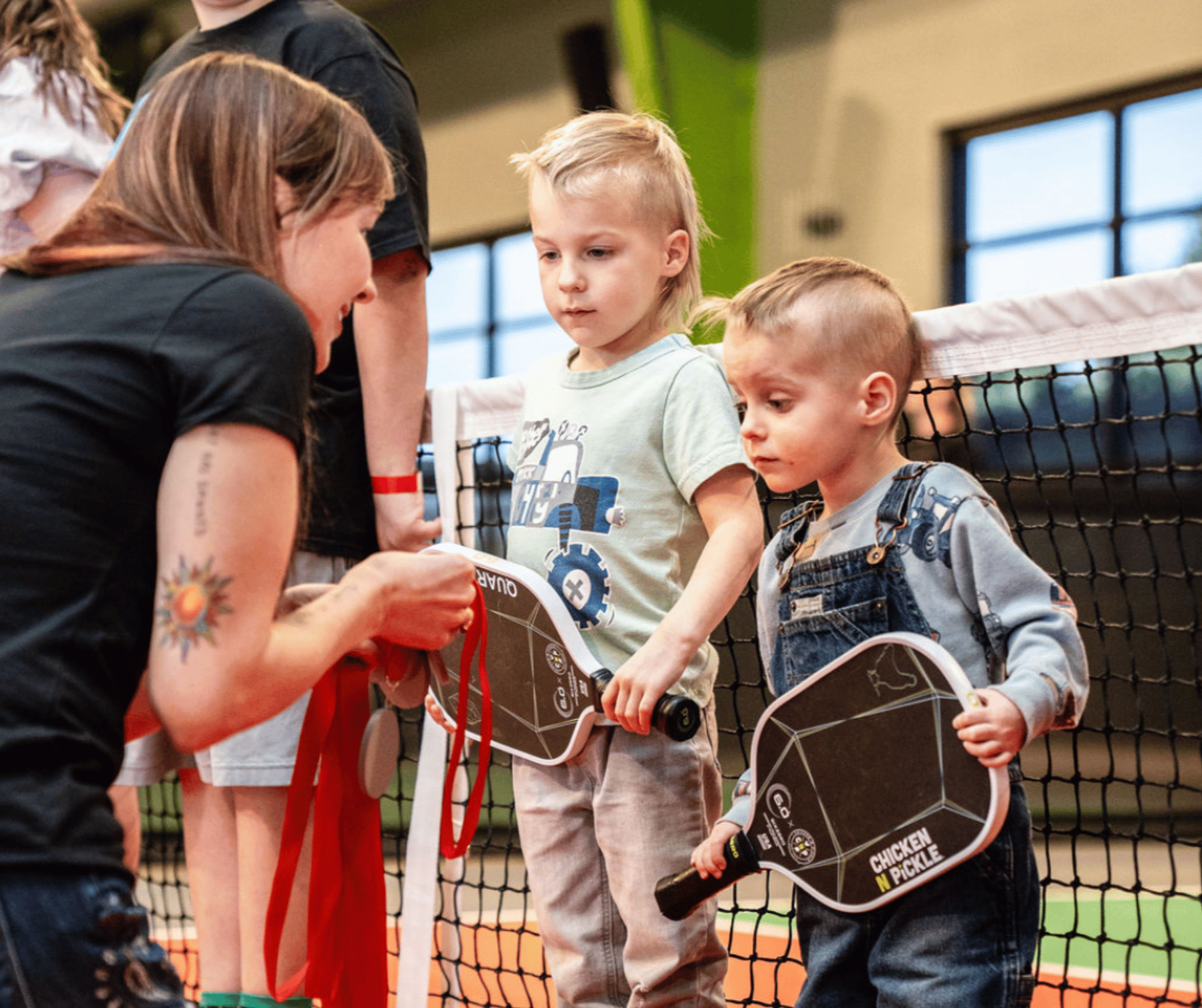 Two small kids holding pickleball paddles and standing in front of a pickleball net look intently at a medal being handed to them by a woman.