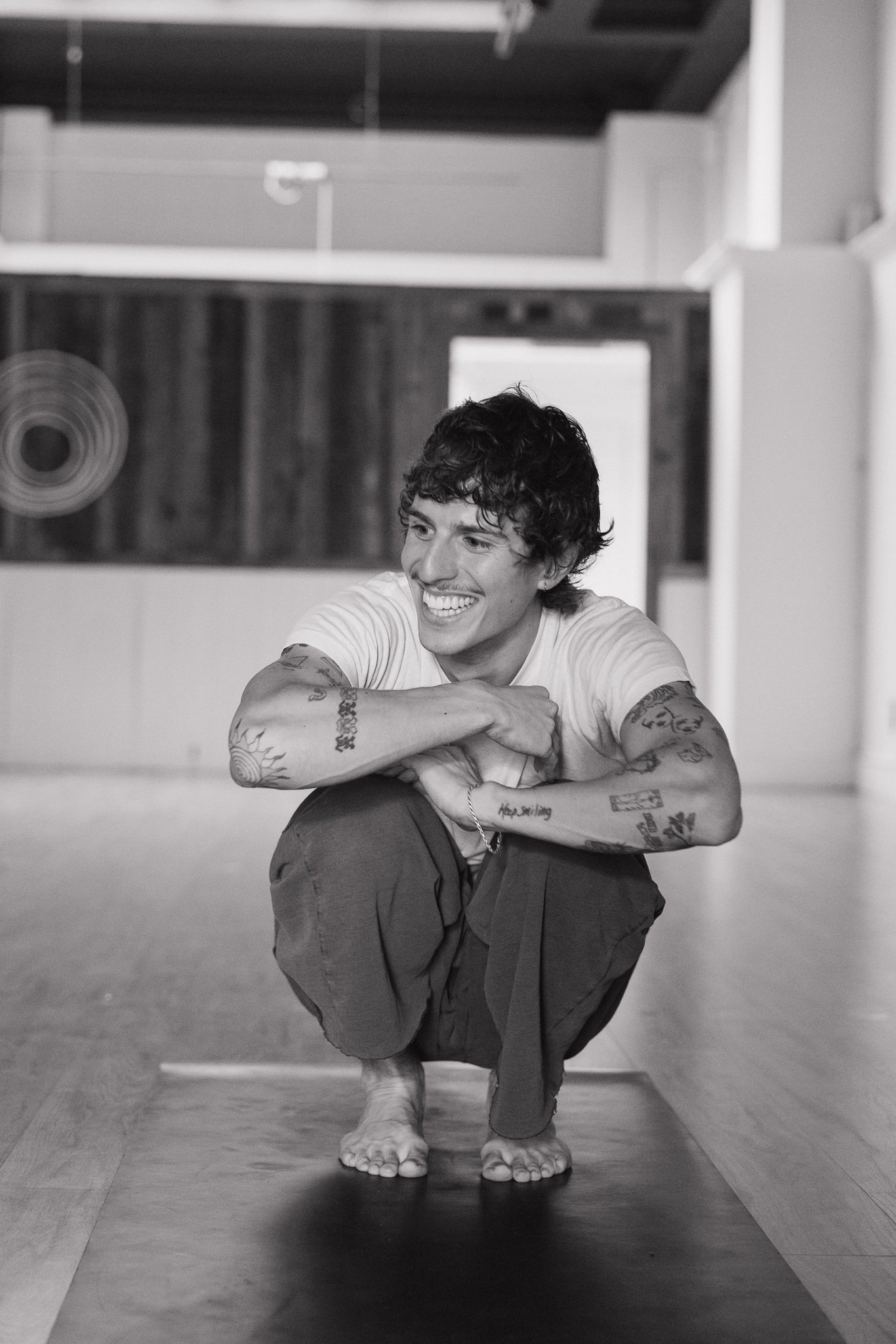 A young man with tattoos smiling while balancing on a yoga mat in a dance studio.