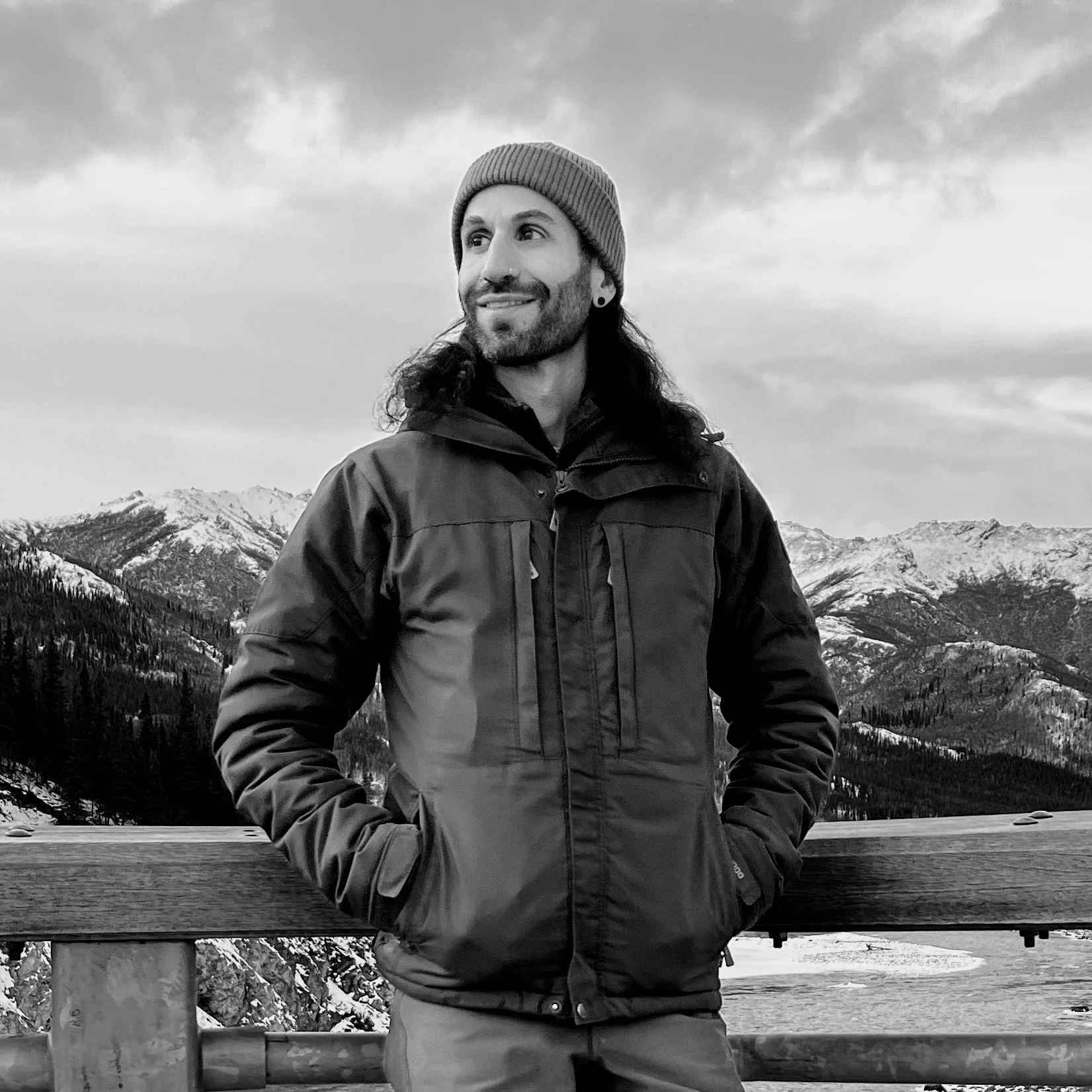 A man with long hair, beard, and nose piercing wearing a beanie and winter jacket, standing outdoors with mountains and cloudy sky in the background.