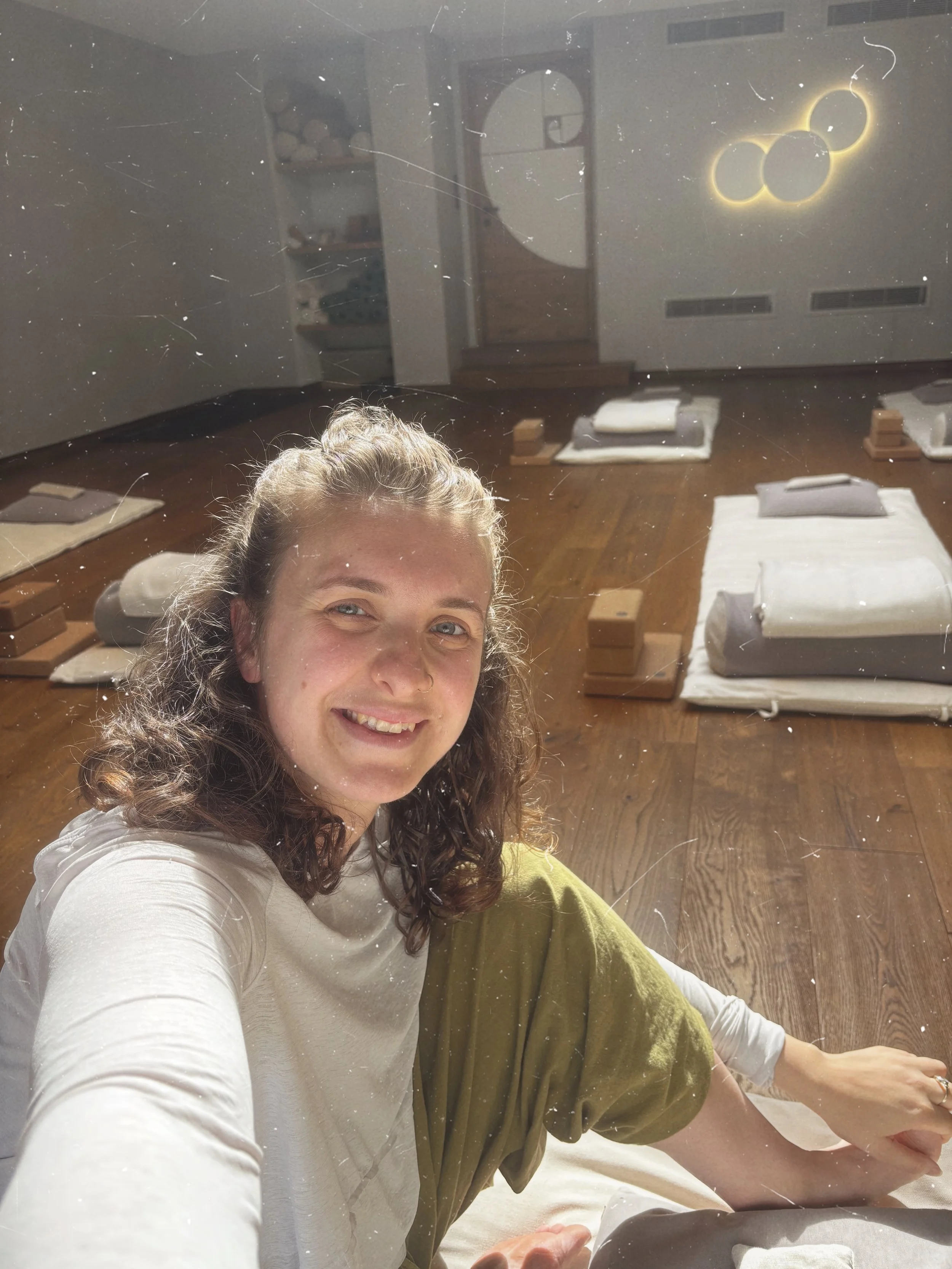 Smiling yoga teacher taking a selfie in a yoga studio with a wooden floor, soft yoga mats, and glowing wall decoration