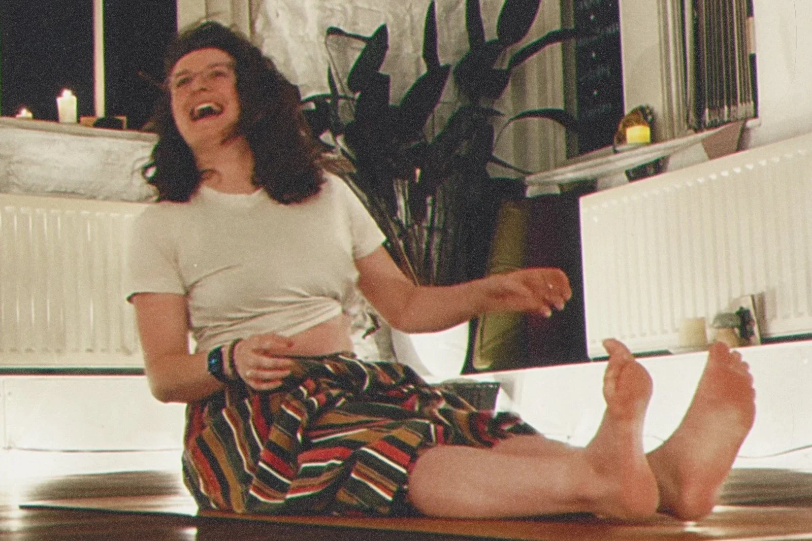 Woman sitting on the floor laughing with her legs outstretched, wearing a white t-shirt and striped pants, in a room with a large plant and a radiator.