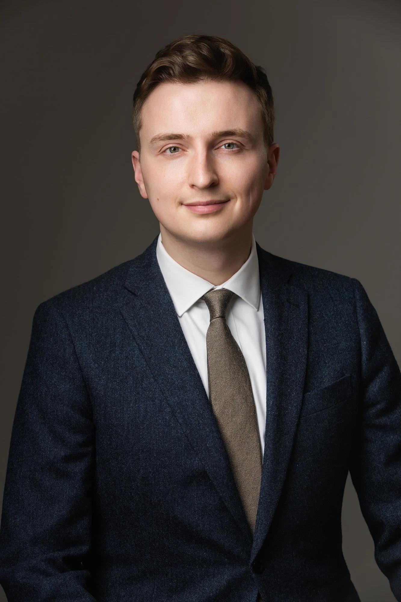 Portrait of a young man with brown hair, wearing a white shirt, brown tie, and dark blazer, smiling slightly against a plain gray background.