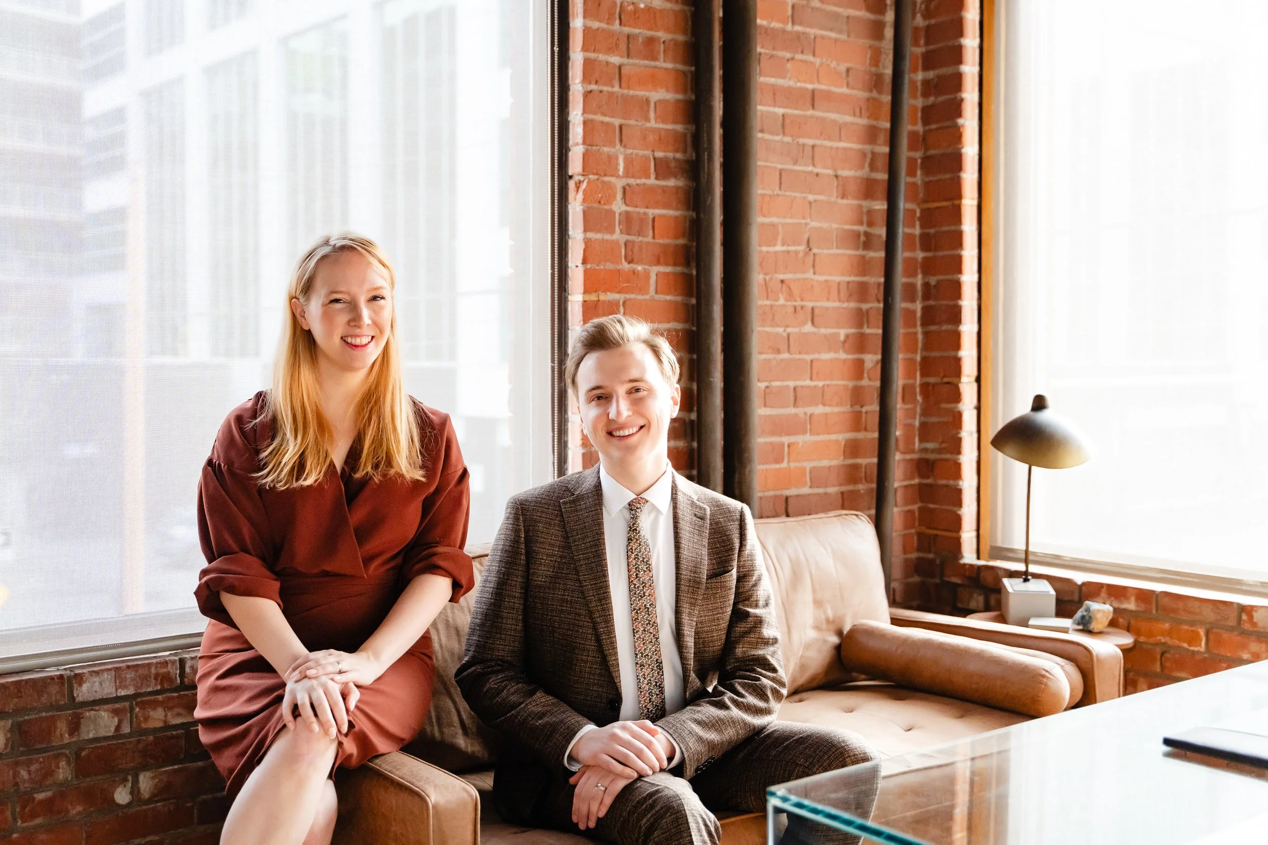 Two smiling young professionals, a woman with long blonde hair wearing a maroon dress and a man with short hair wearing a brown plaid suit, sitting in a modern office with brick walls and large windows, during the daytime.