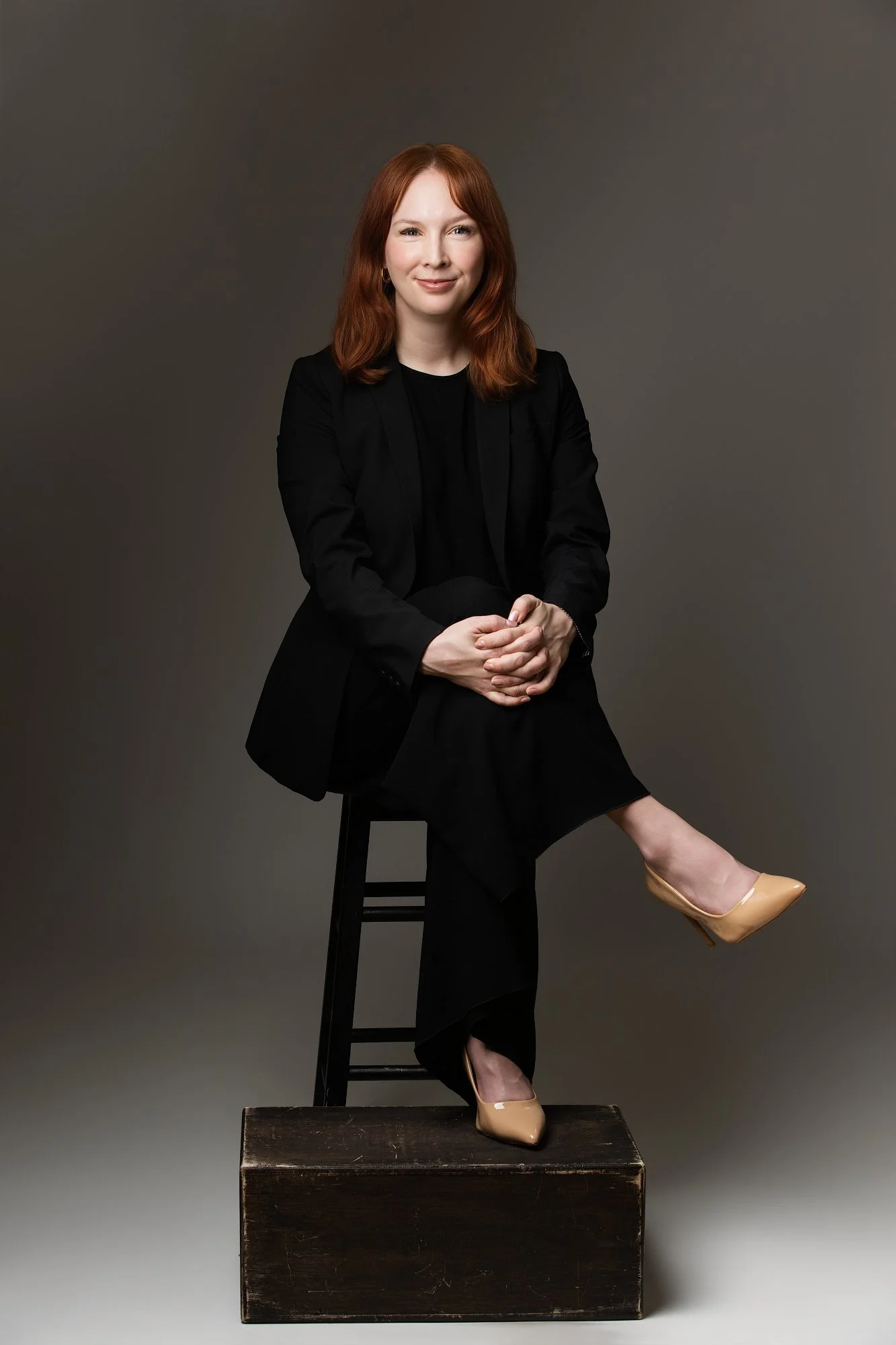 A woman with red hair, dressed in black, sitting on a stool with one leg crossed over the other, wearing beige high heels, against a plain gray background.
