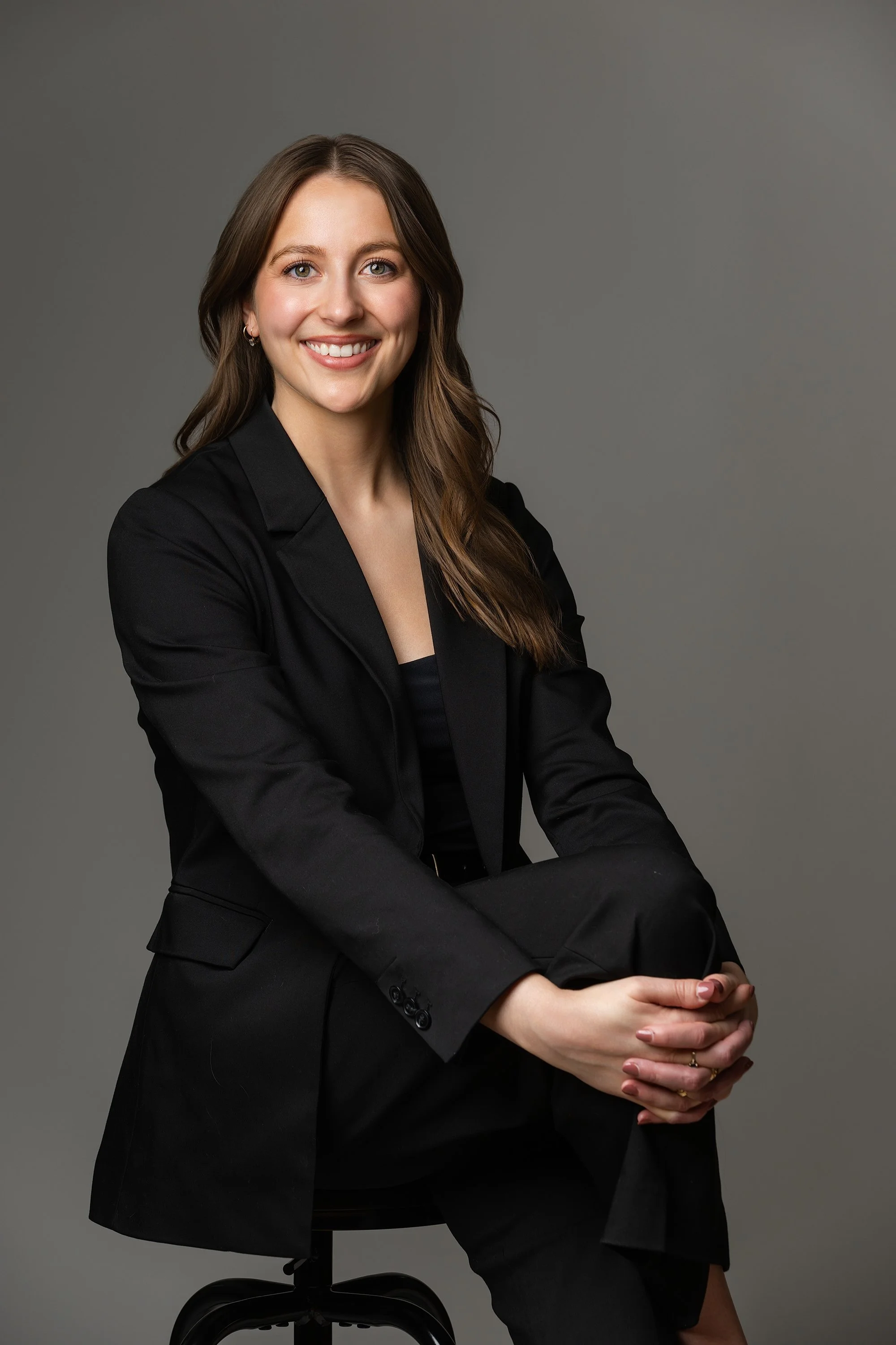 A woman with long brown hair, wearing a black blazer and black pants, sitting on a black office chair against a plain grey background, smiling and clasping her hands.