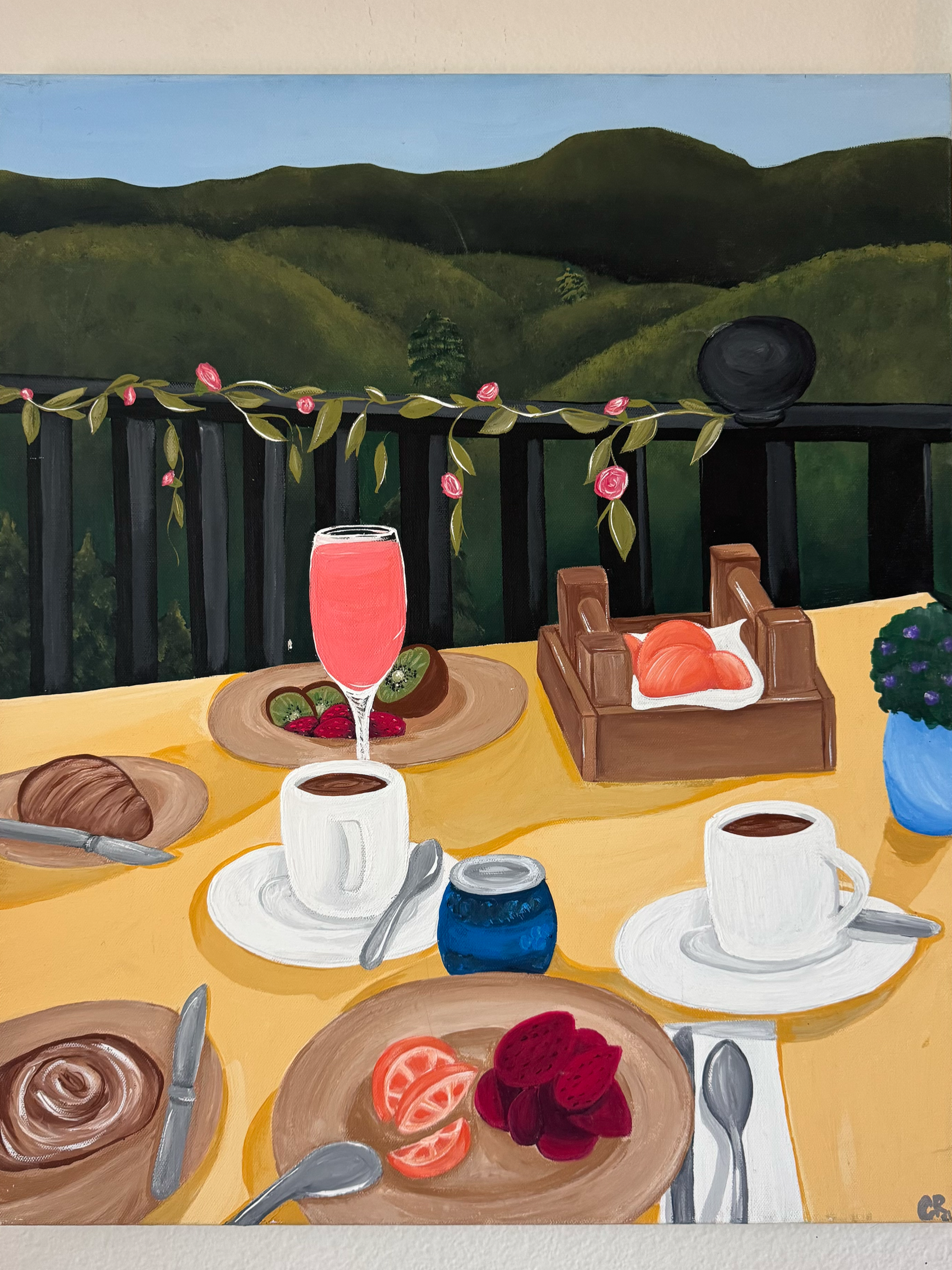 A painting of an outdoor breakfast scene on a yellow table with a mountain view in the background. The table has cups of coffee, plates with fruit and pastries, a glass of pink juice, and a flower pot with purple flowers. There is also a tray with ad