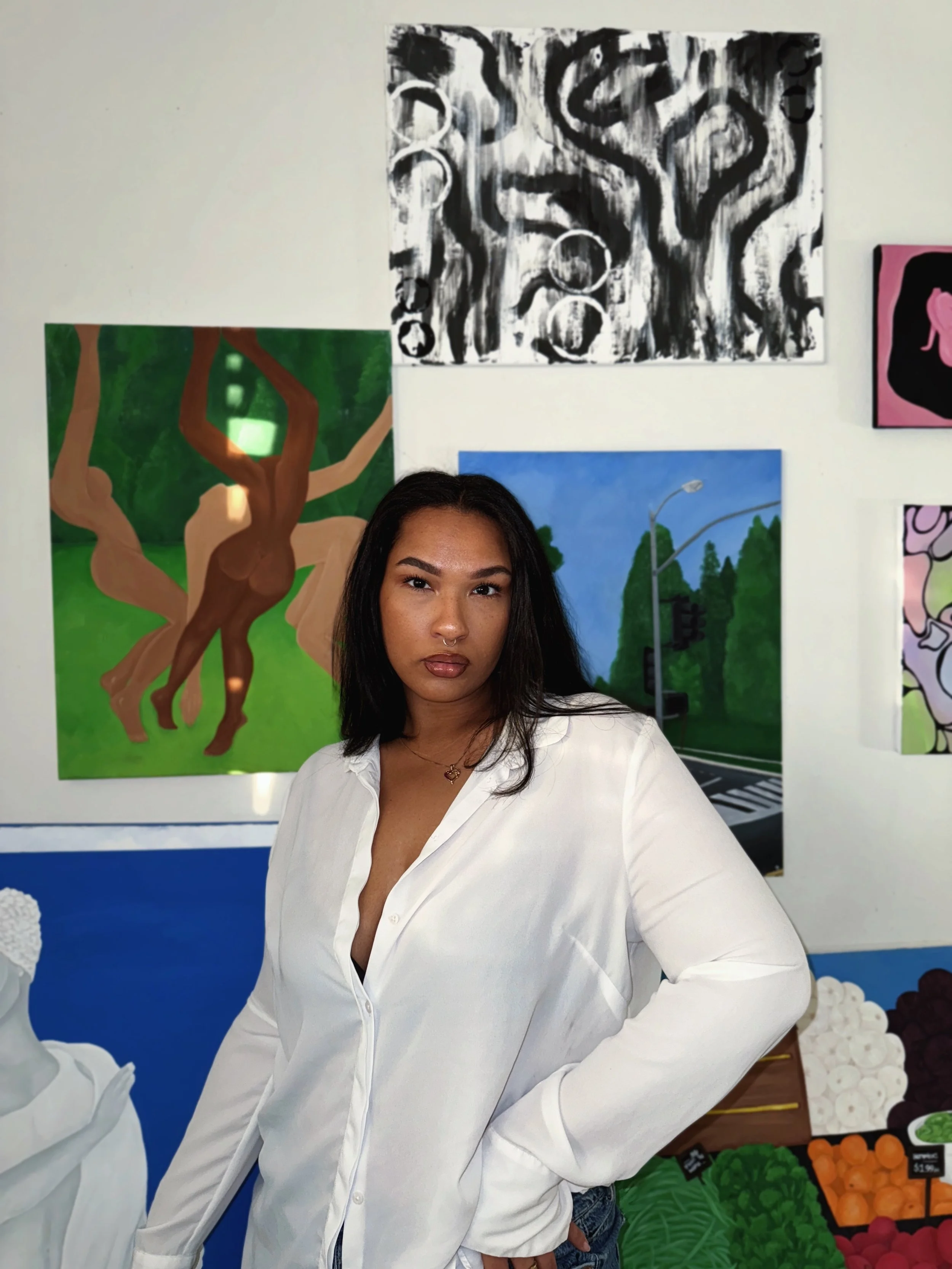 A young woman with dark hair wearing a white button-up shirt standing in front of colorful paintings on an art gallery wall.