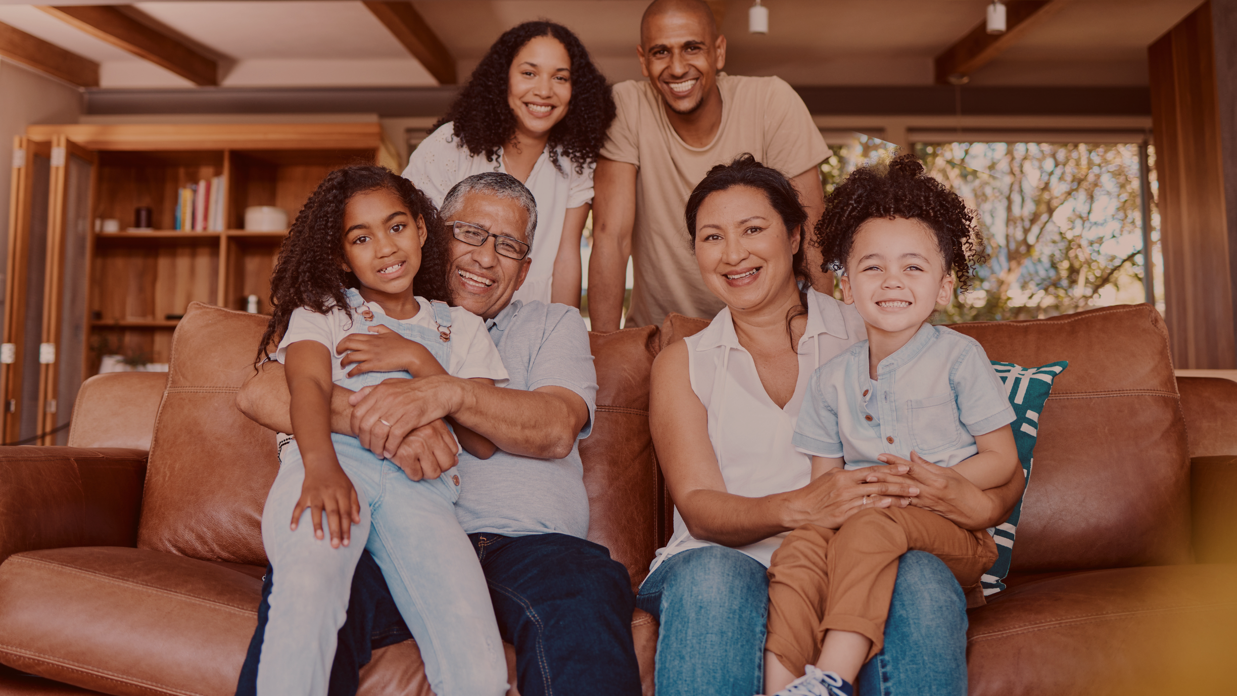 Multi-generational family smiling and sitting on a leather sofa in a cozy living room with wooden ceiling beams, bookshelves, and large windows showing outdoor trees.