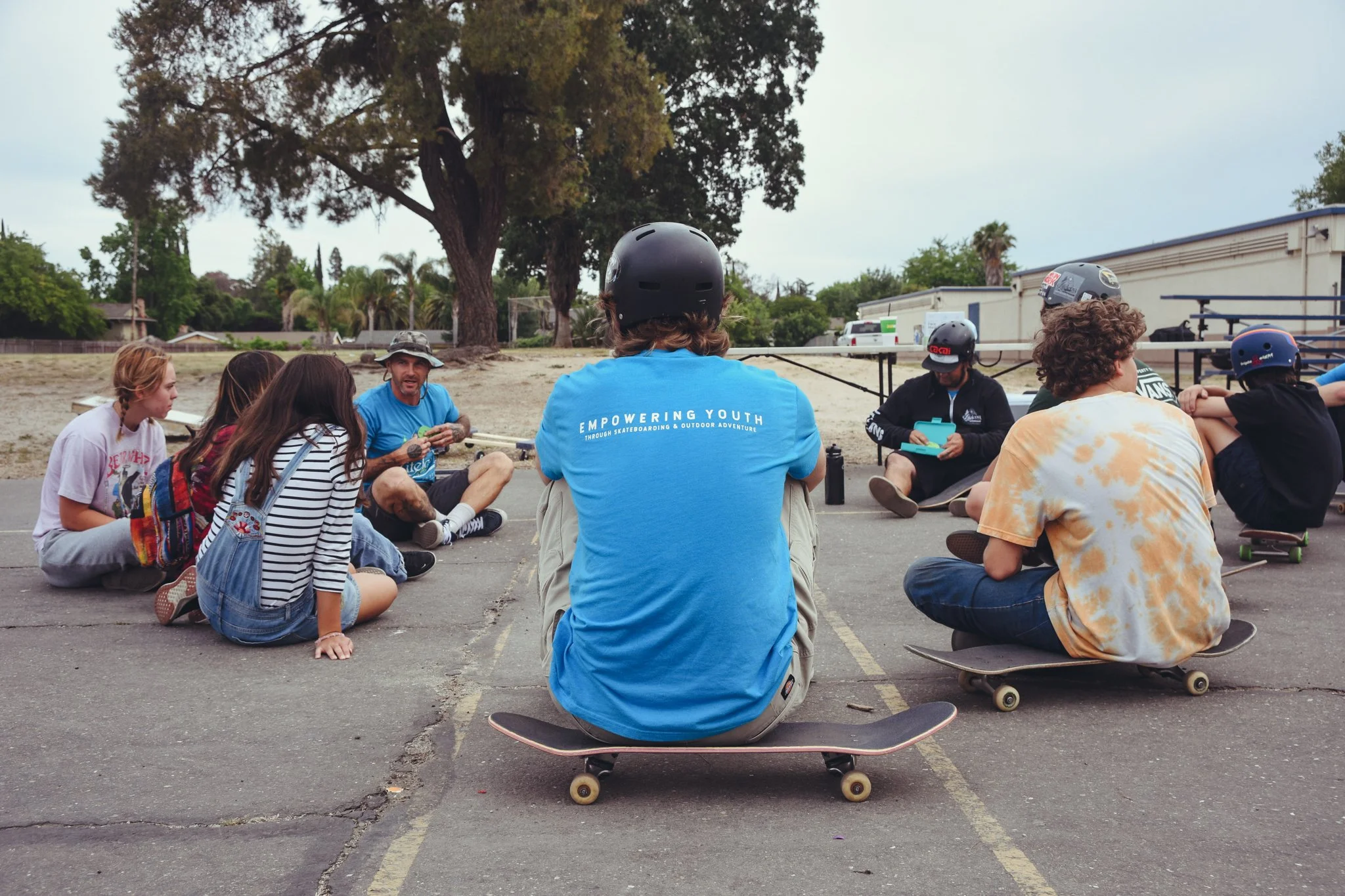 Group of young people sitting on skateboards on an asphalt surface in a park, listening to a skateboarding instructor. Some are wearing helmets, and there are trees and a building in the background.