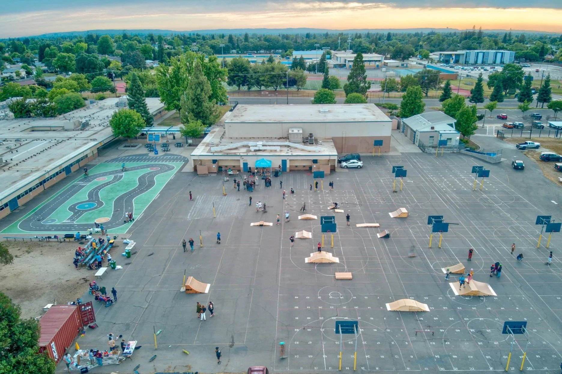 Aerial view of a school parking lot with children and adults gathering, a painted playground with pathways, and a large school building surrounded by trees.