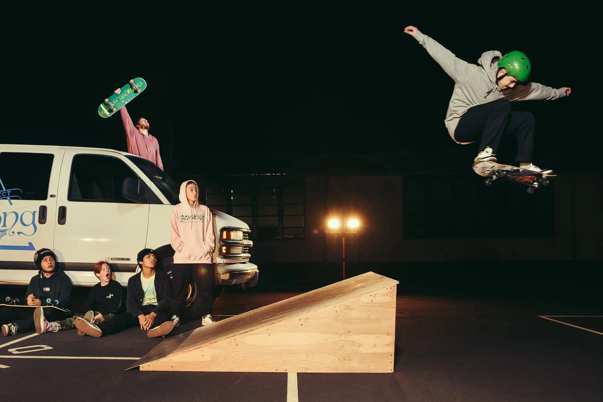Skateboarder in green helmet performing a jump above a ramp at night, with a group of young people sitting and standing nearby, some with skateboards, in a parking lot illuminated by streetlights.