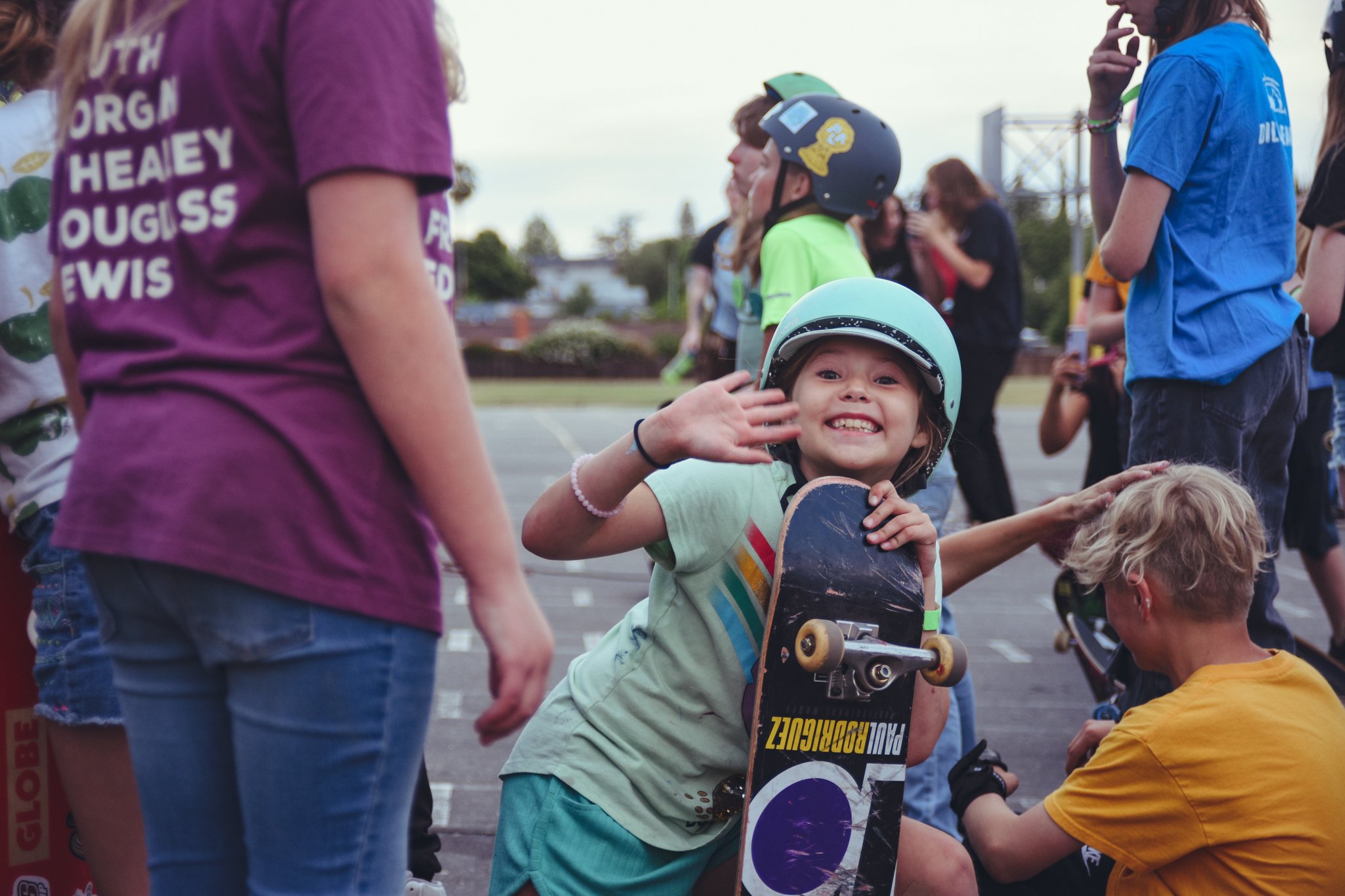 A young girl smiling and waving, wearing a helmet and holding a skateboard, surrounded by people outdoors during the daytime.