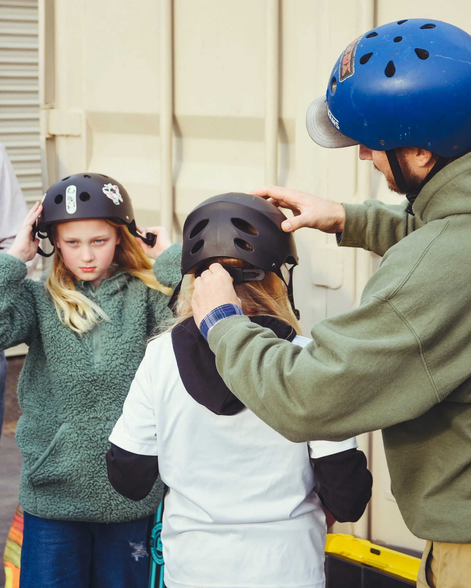 An adult helping a girl put on a black helmet, with another girl waiting nearby, both wearing helmets, in an outdoor setting.