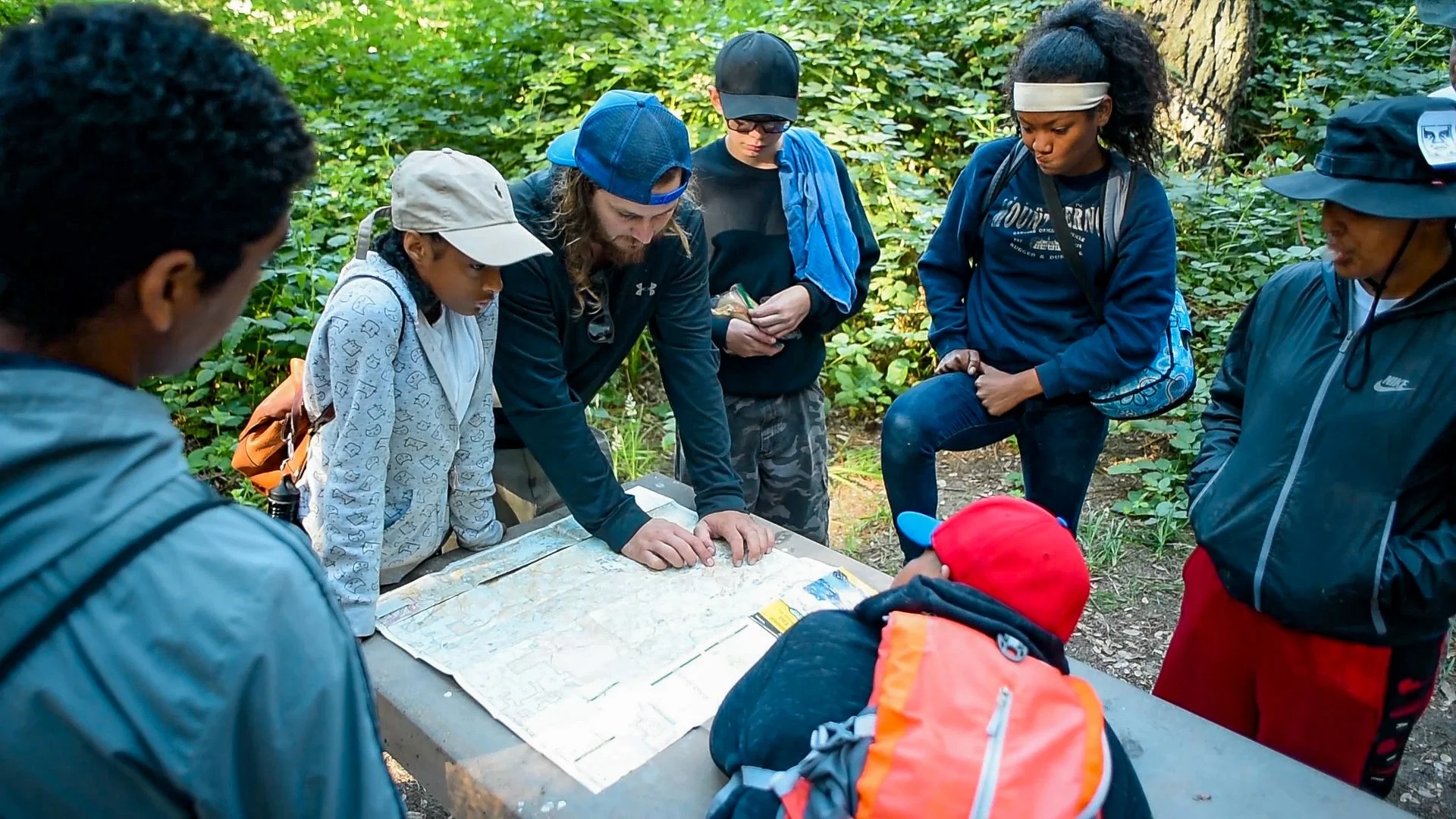 Group of people gathered around a map on a wooden table outdoors in a forested area.
