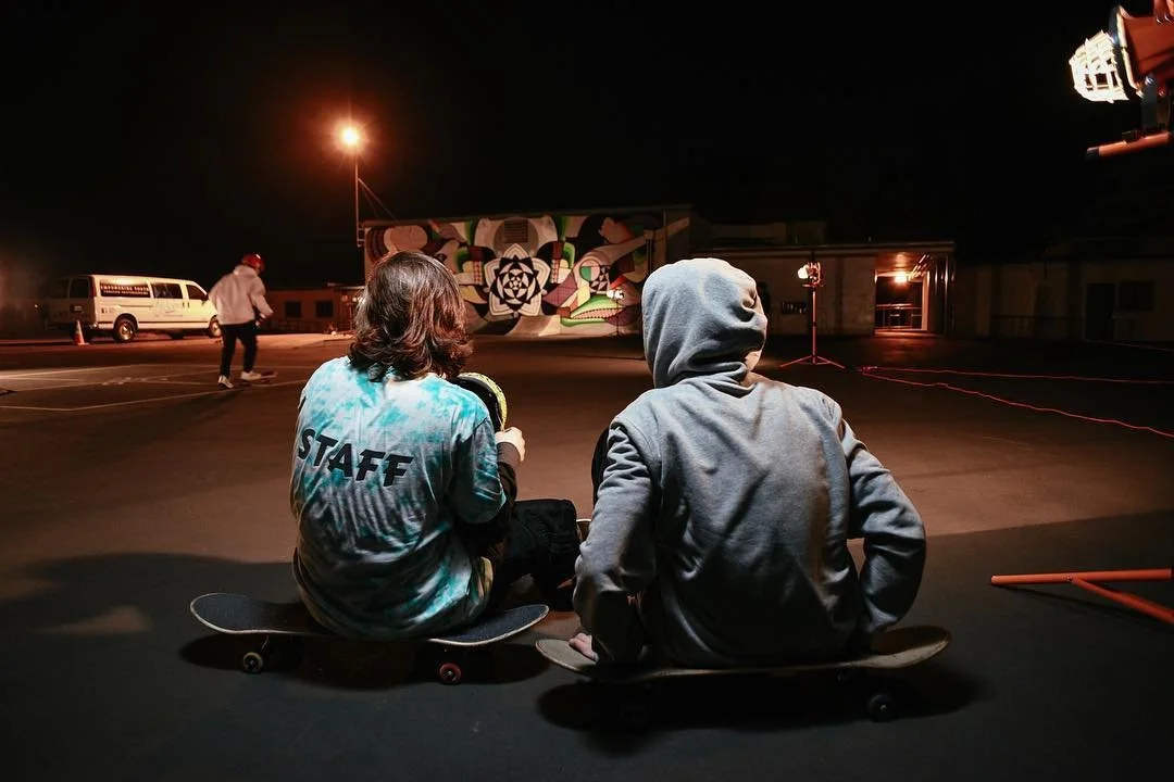 Two skateboarders sitting on the ground at night, with one wearing a hoodie and the other a staff shirt, watching a person skateboarding in a parking lot illuminated by streetlights and colorful wall art in the background.