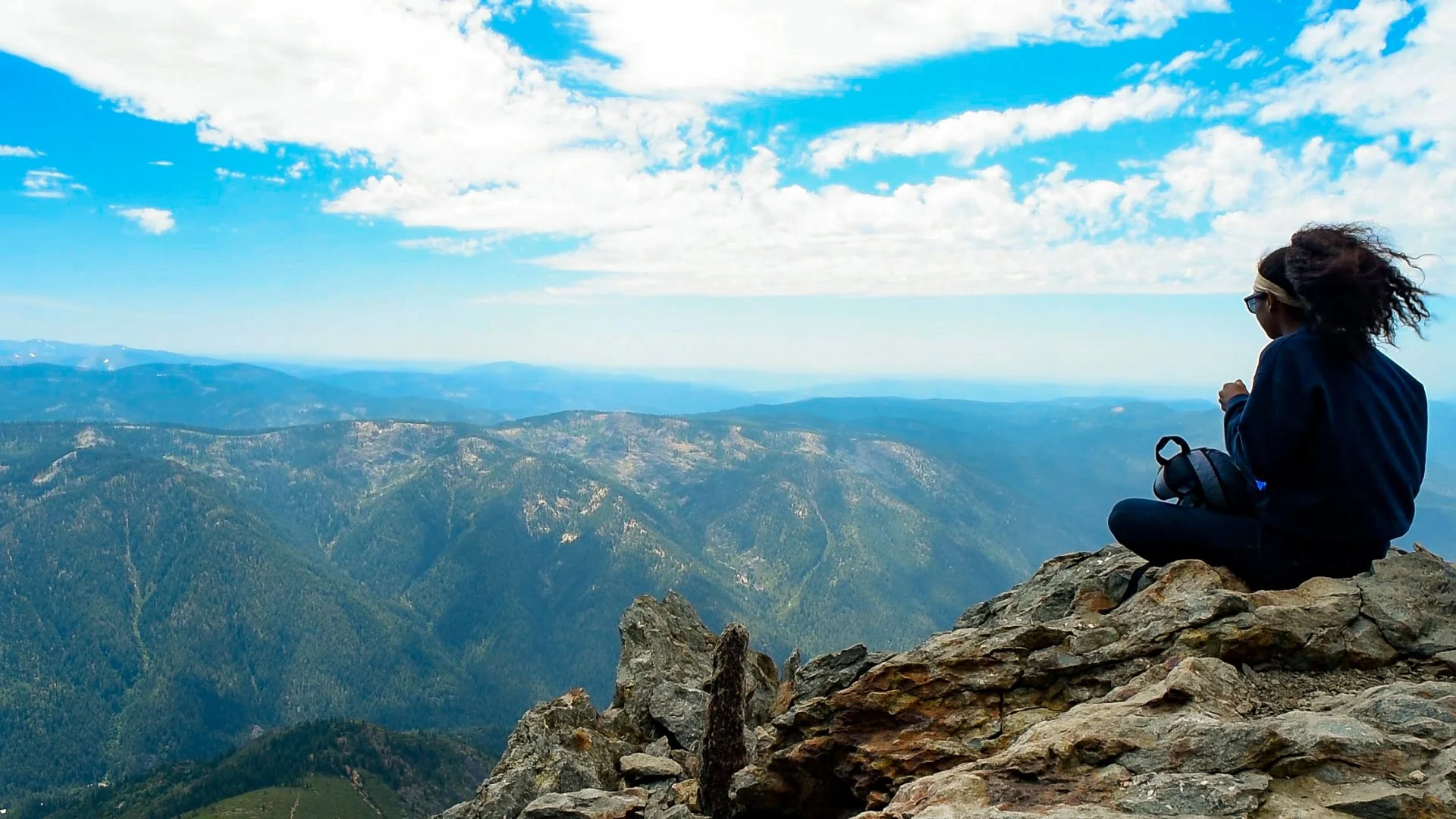 A woman sitting cross-legged on a rocky mountain edge, overlooking expansive mountain ranges and valleys under a partly cloudy sky.