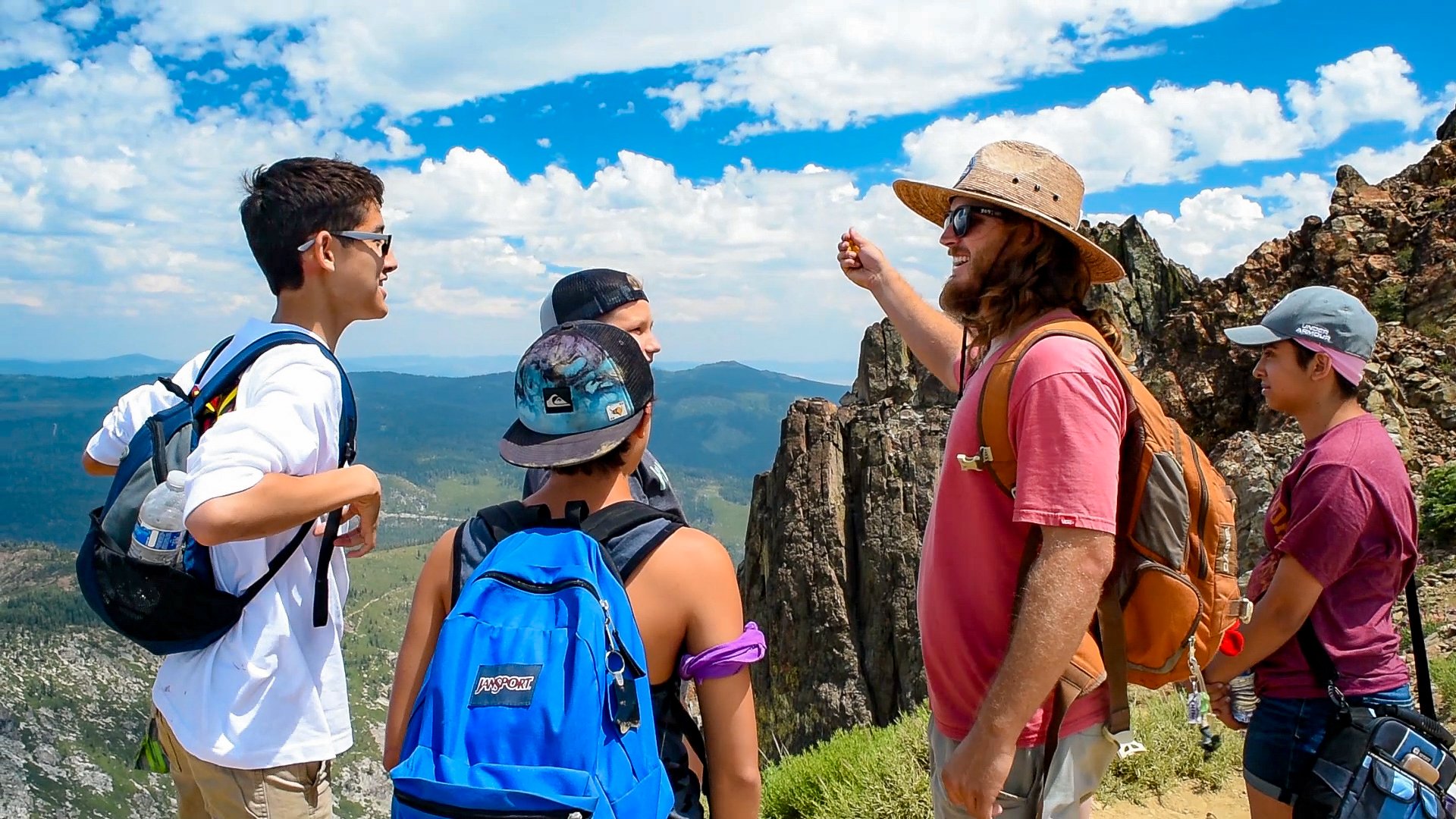 A group of people hiking outdoors, standing on a trail with rocks and green grass, with a scenic mountain view and a blue sky with clouds in the background. One person appears to be giving instructions or talking to the others.