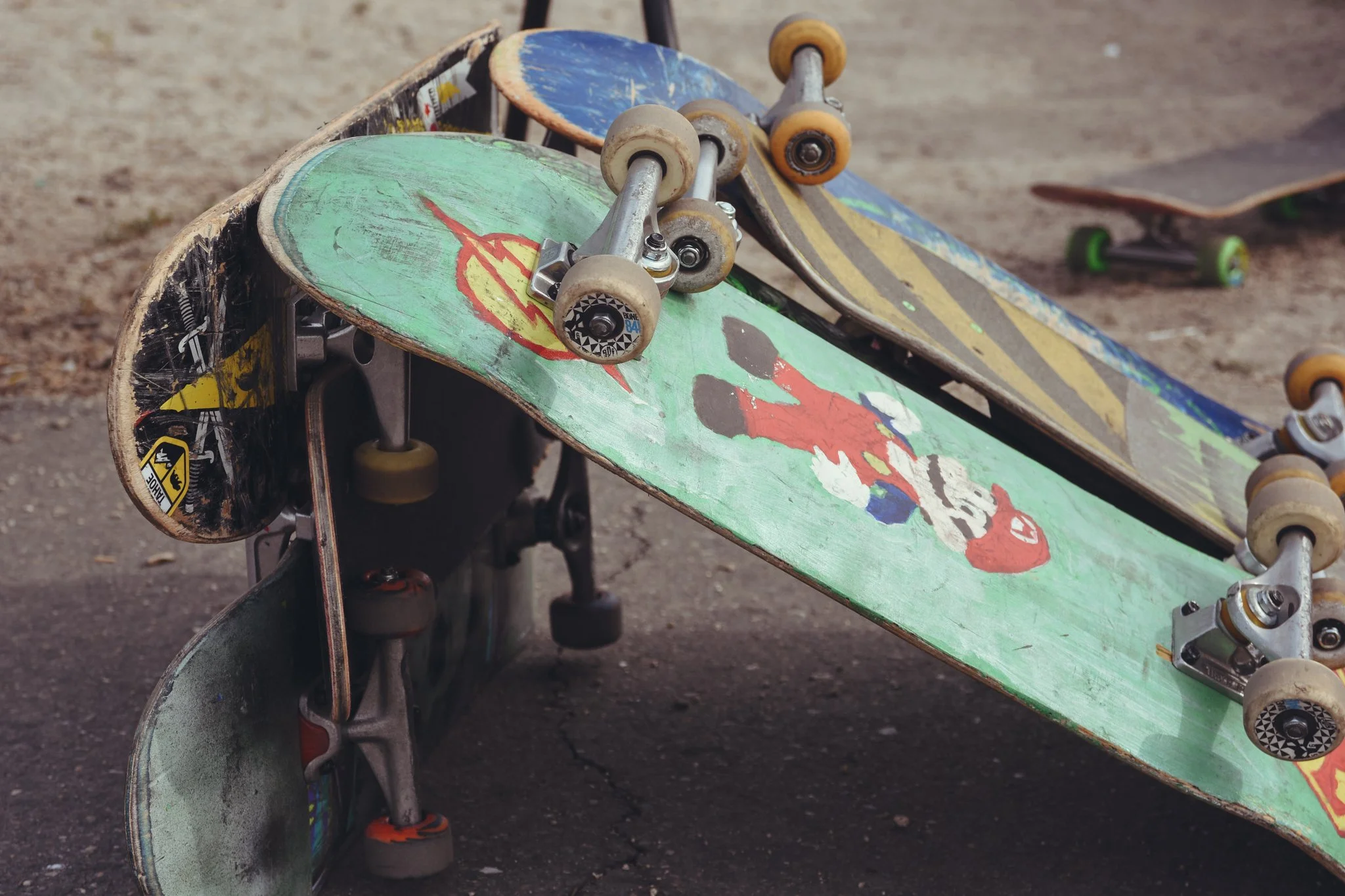 Skateboards stacked on a rough ground with worn wheels and painted decks, one deck featuring a colorful cartoon character.