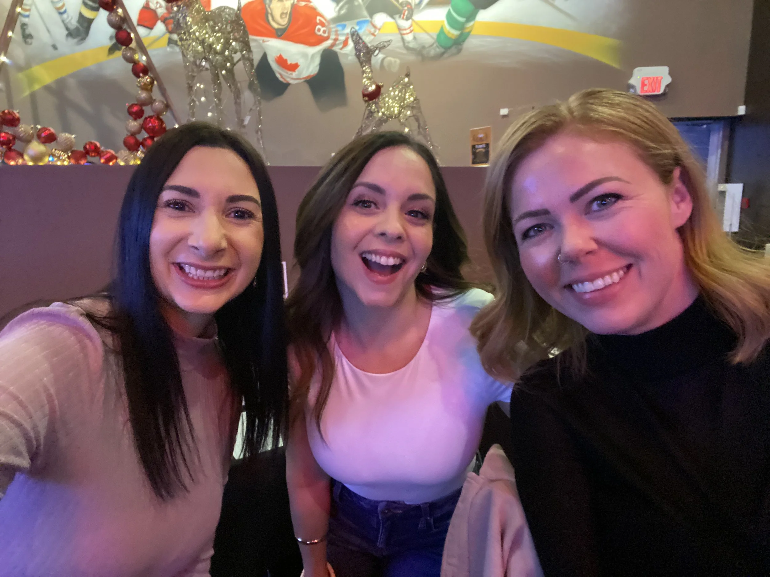 Three women smiling and posing for a selfie in a decorated indoor setting with Christmas ornaments.