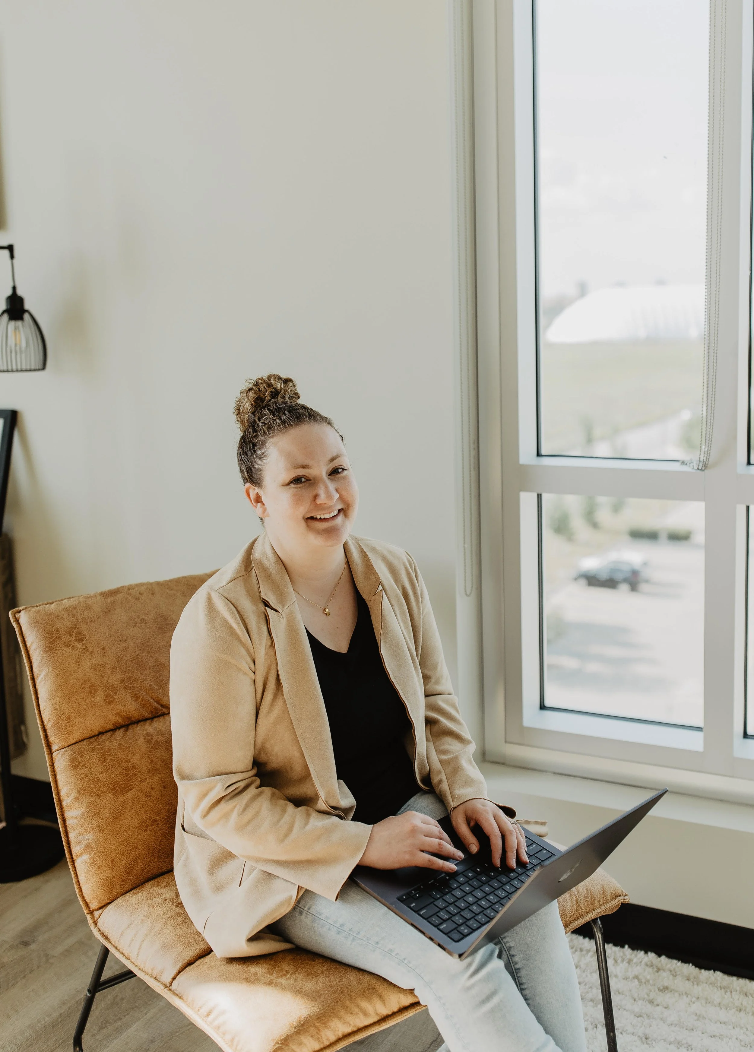 Smiling woman sitting on a beige chair working on a laptop by a large window in a bright room.