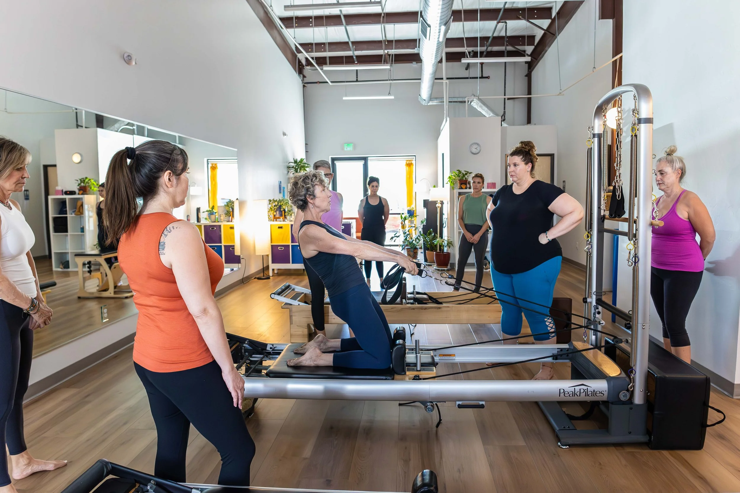 A group of women participating in a Pilates class using a reformer machine in a bright, modern fitness studio.