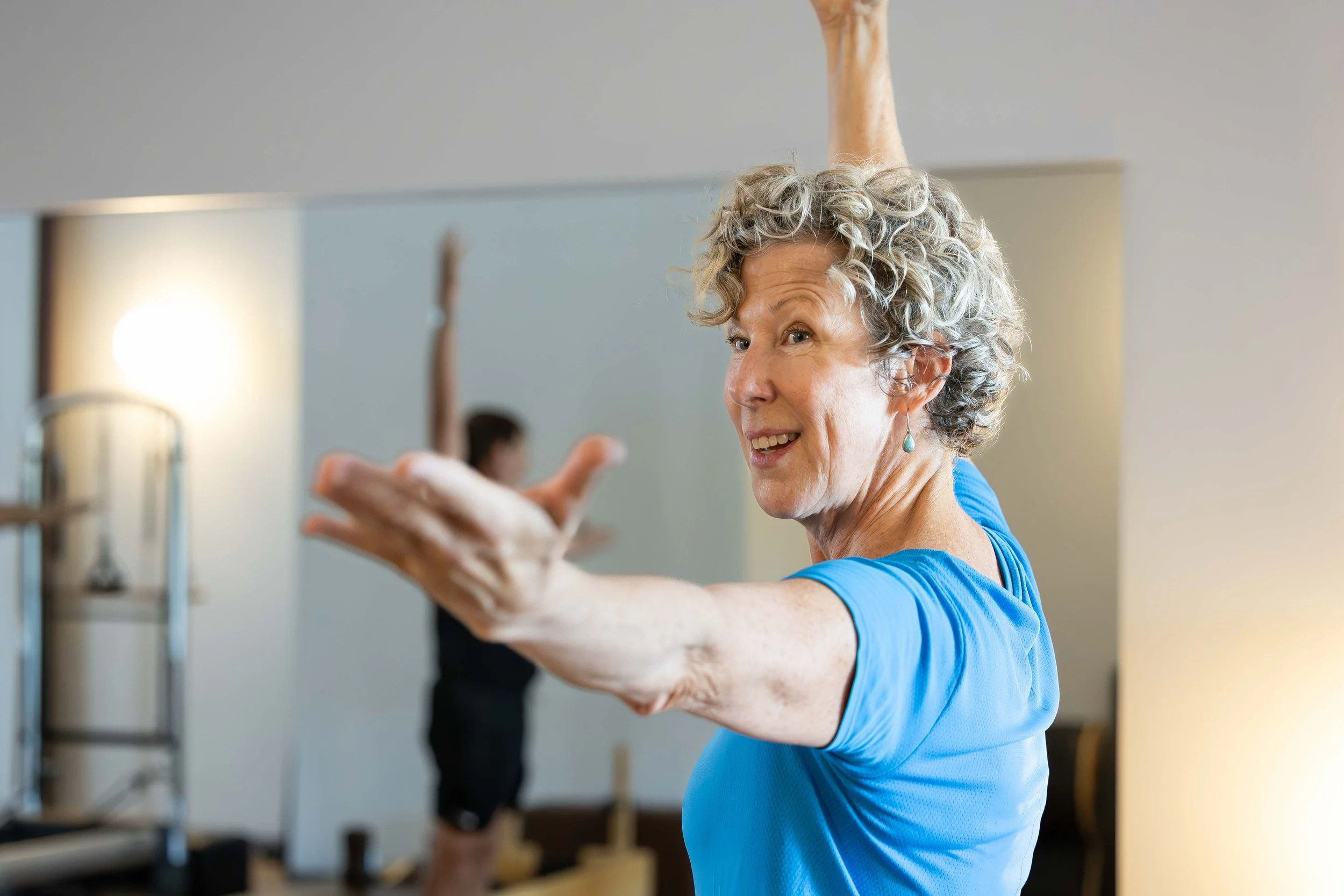 An elderly woman with curly gray hair and earrings is practicing a stretch or yoga pose in a fitness or dance studio, with her arms extended and smiling.