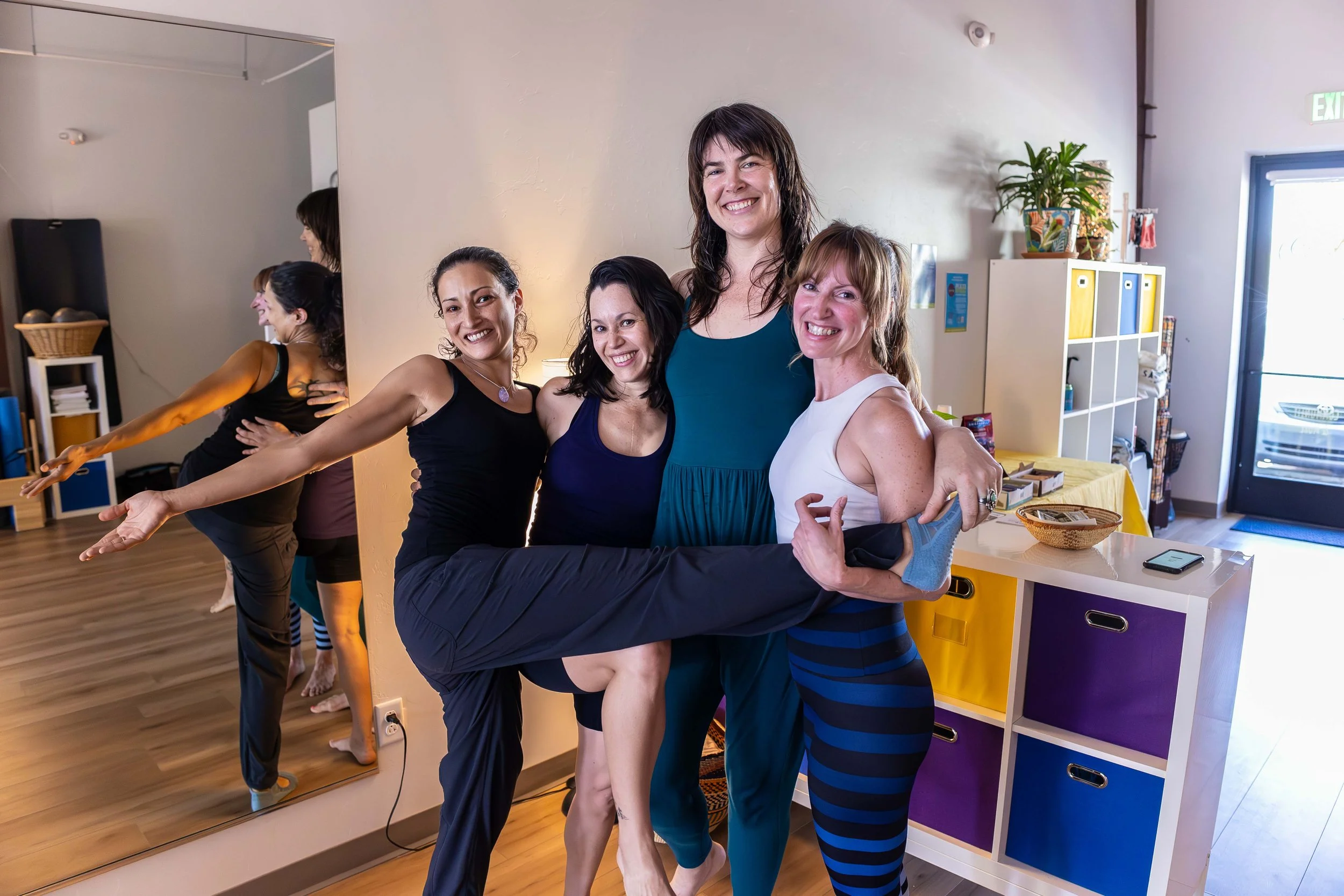 A group of five women in a dance studio, with one woman doing a leg kick and all of them smiling and posing together affectionately.