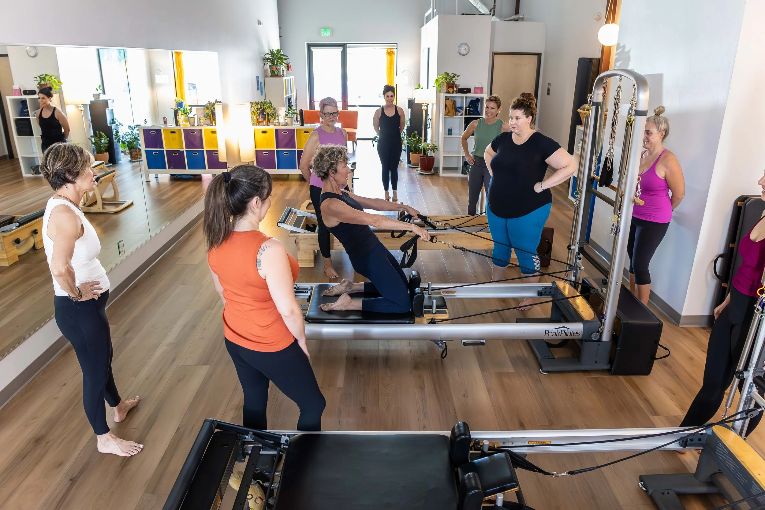 Group of women participating in a Pilates class using specialized reformer machines in a bright studio with wooden floors and potted plants.